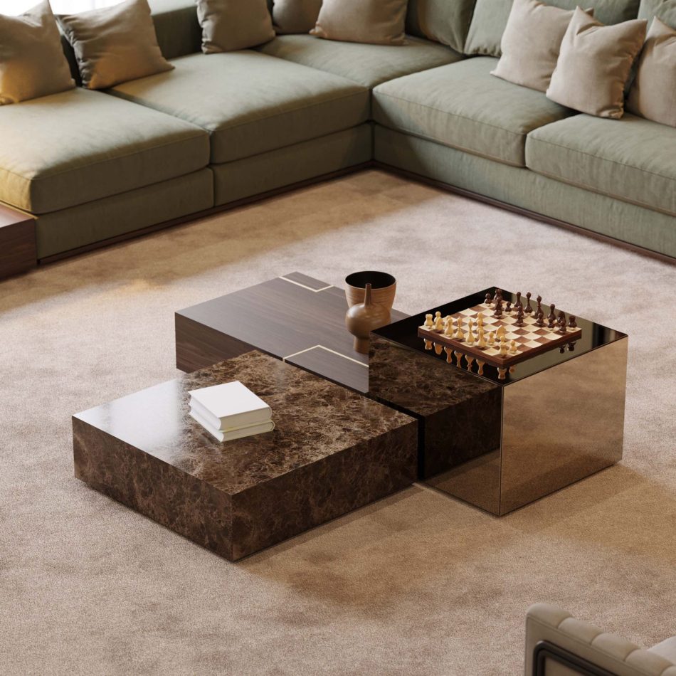 The Fortune Coffee Table trio, in dark marble, warm wood, and reflective metal, stands before a green sectional with beige cushions. Decorative books, a wooden vase, and a chessboard add texture to the soft, neutral room. The setting highlights sculptural contemporary furniture, refined design details, and the crafted presence of high end furniture.