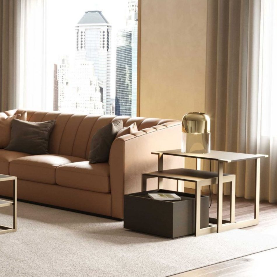 The Fortune Side Table, with its brushed bronze‑brass frame and high‑gloss dark oak surfaces, sits beside a tan leather sofa with soft grey cushions. A gold lamp rests on the upper tier, framed by floor‑to‑ceiling curtains and an urban skyline beyond the window. The setting blends warm living room furniture with sculptural modern furniture and a calm sense of interior design inspiration.