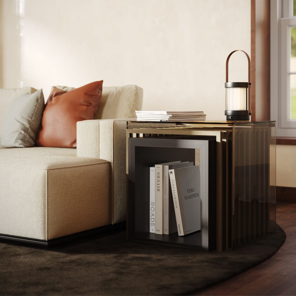 The His Side Table appears beside a beige sofa, revealing its three stacked cubic surfaces finished in bronze‑toned materials. Styled with books and a lantern‑style lamp, the piece blends modern furniture, warm design details, and elegant interior design inspiration.