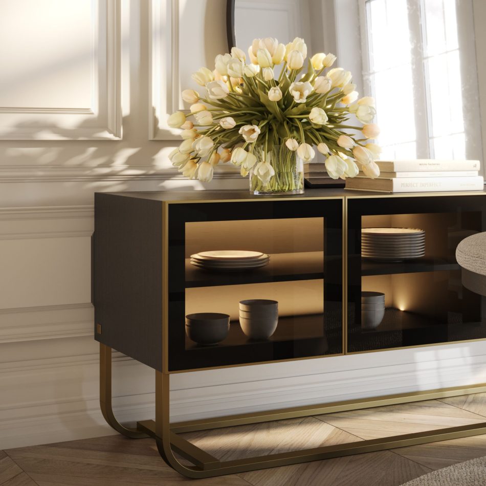 The His Sideboard features four bronze‑glass doors illuminated from within, showcasing neatly arranged dining pieces with a soft golden glow. A tall vase filled with pale tulips and a small stack of books rest above the sideboard, blending luxury dining room furniture, warm design details, and elegant interior design inspiration.