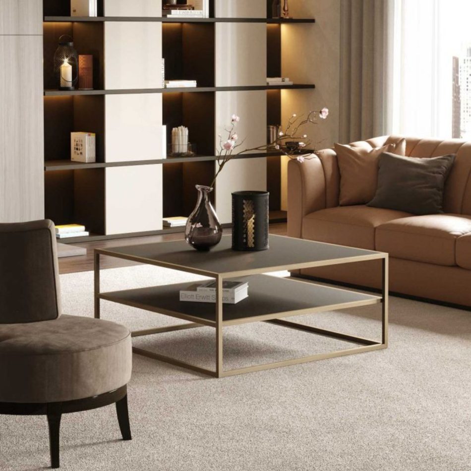 The Bridge Coffee Table, featuring a metallic frame and two dark shelves, stands before a tan leather sofa and a softly illuminated shelving wall. A glass vase with branches and curated décor add texture to the warm, neutral setting. The room blends elegant high end furniture with sculptural casegoods hospitality and a calm, contemporary living room design.