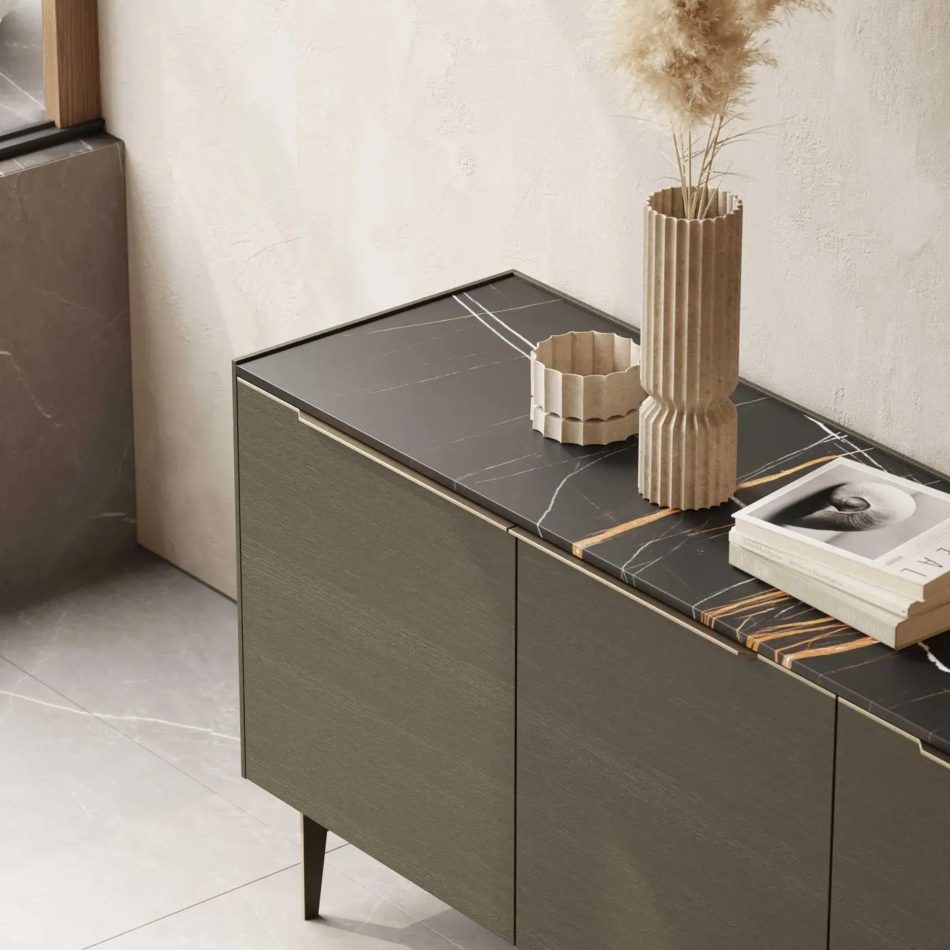 The Landform Sideboard stands on slender legs, its dark marble top accented by soft white and gold veining. A ribbed beige vase with dried pampas and curated objects introduce warmth against the textured wall and polished floor. This calm vignette highlights the precision of modern furniture, the crafted presence of luxurious furniture, and the sculptural harmony found in thoughtful design details.