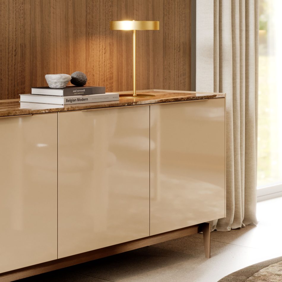 A glossy beige sideboard with a marble top is styled with stacked books, stone objects, and a sculptural gold lamp, set against warm wood panels and soft curtains. Natural light enhances the calm, minimalist composition, reflecting the refinement of luxury home furniture, the clean lines of modern furniture, and the quiet elegance found in thoughtful design details.