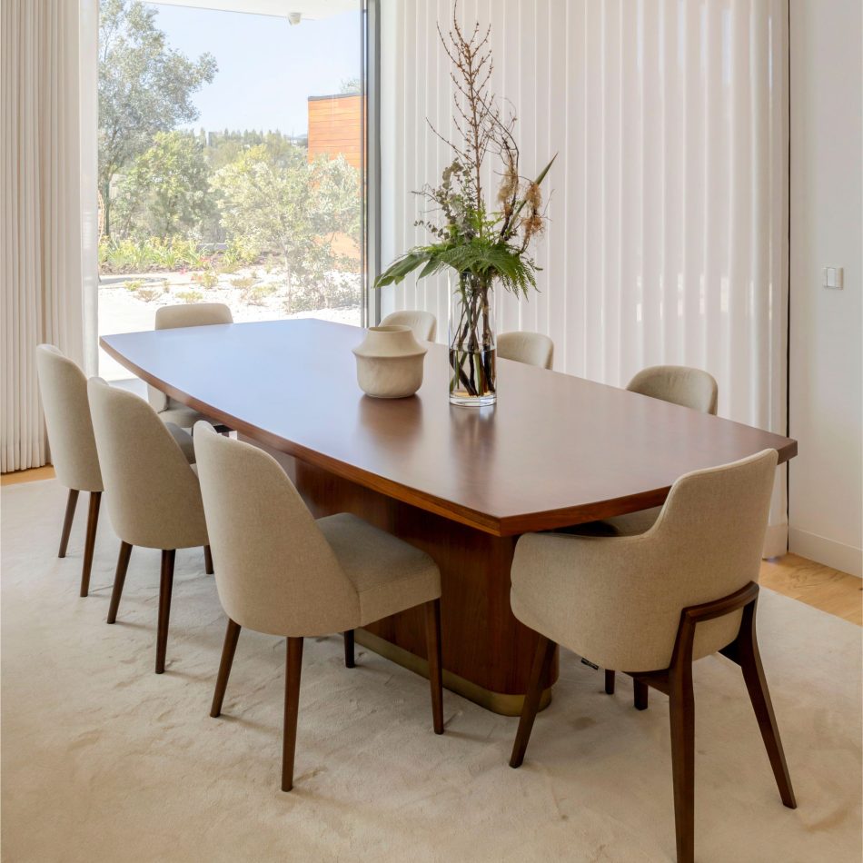 The Elea Dining Table extends across a bright dining room, its smooth oak surface paired with bronze‑lacquered detailing that adds a subtle touch of luxury. Surrounded by eight light‑beige upholstered chairs, the table’s generous proportions highlight its ability to host large gatherings with ease. Natural light, clean lines and warm materials enhance the atmosphere, expressing the balance of modern furniture design and the durability of contemporary dining furniture.