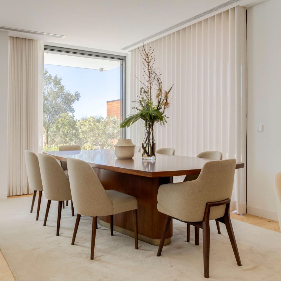 The Elea Dining Table in oak wood stands at the centre of a serene dining room, in a layout designed for comfort and functionality. A tall glass vase with green branches adds height and freshness, while soft natural light filters through floor‑to‑ceiling windows. The scene highlights the table’s firm structure, elegant bronze detailing and its role as a statement piece within dining room interiors.
