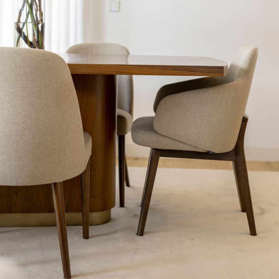 A close view of the Elea Dining Table reveals its smooth rectangular oak top and rounded edges, paired with upholstered beige chairs that echo its warm tones. The composition emphasises the table’s solid construction, contemporary silhouette and refined materiality. Soft lighting and a neutral backdrop enhance the tactile quality of the wood, underscoring the table’s blend of innovation, functionality and modern furniture design.