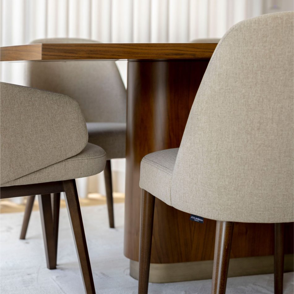 A close view of the Elea Dining Table reveals its smooth oak surface and rounded edges, paired with light beige upholstered chairs that echo its warm tones. Soft curtains and gentle lighting enhance the tactile quality of the wood, highlighting the table’s sturdy construction and the simplicity of modern furniture design.