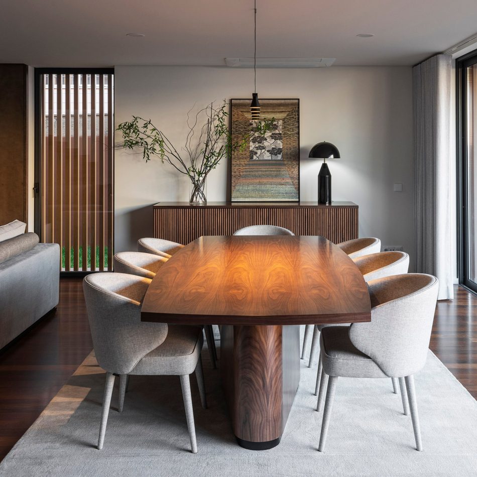 The Elea Dining Table stands at the centre of a contemporary dining room, its polished oak top and bronze‑detailed base complemented by six curved upholstered chairs. A wooden sideboard with vertical slats, a sculptural lamp and a tall vase of green branches frame the scene, creating a warm, elegant setting that reflects the charm and quality of contemporary dining furniture.