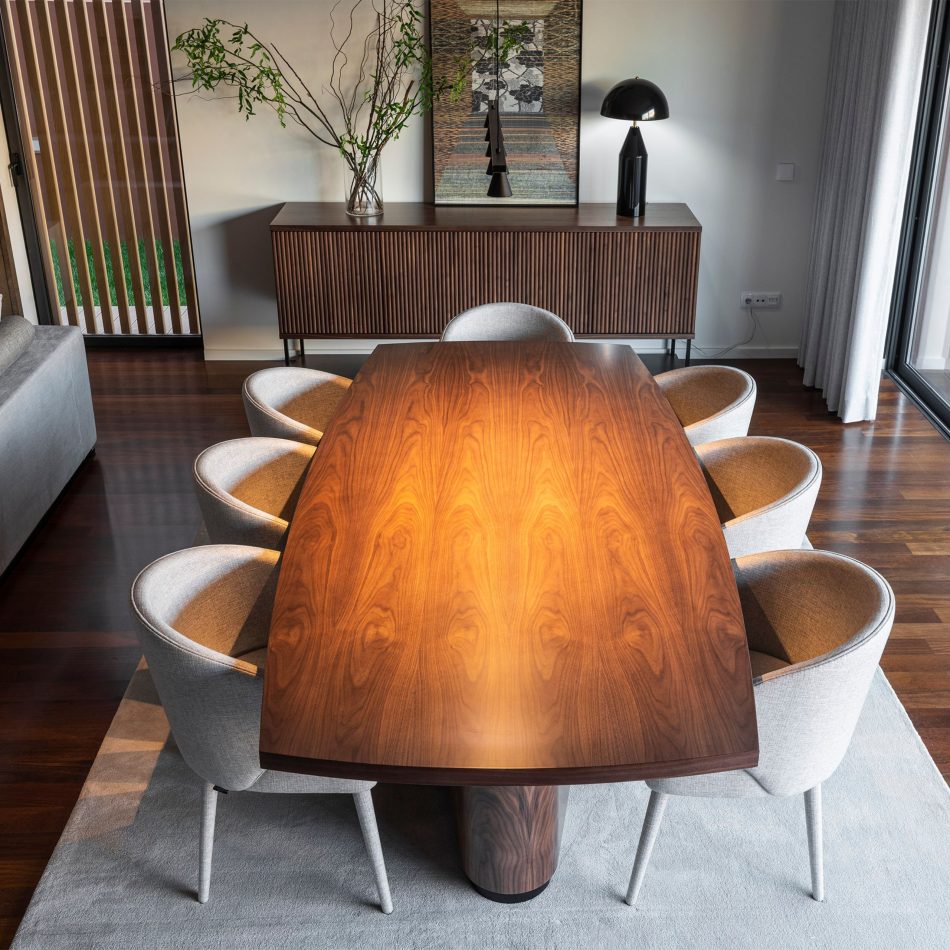 The Elea Dining Table with a rich wood‑grain surface anchors the dining room, surrounded by six light upholstered chairs that enhance its generous proportions. Natural light from tall windows highlights the table’s firm structure and luxurious bronze detailing, reinforcing its role as a functional, inviting centrepiece within dining room interiors.