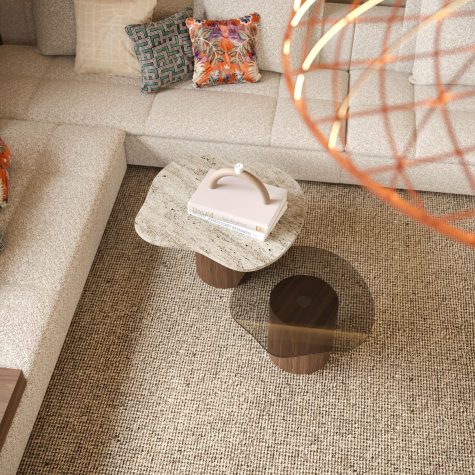 The Natur Side Tables are shown as a pair with rounded tops in stone and dark glass finishes, placed on a woven neutral rug beside a beige sofa with patterned cushions. Styled with a sculptural object and books, the composition reflects modern coffee table design, subtle design details, and furniture luxury design.
