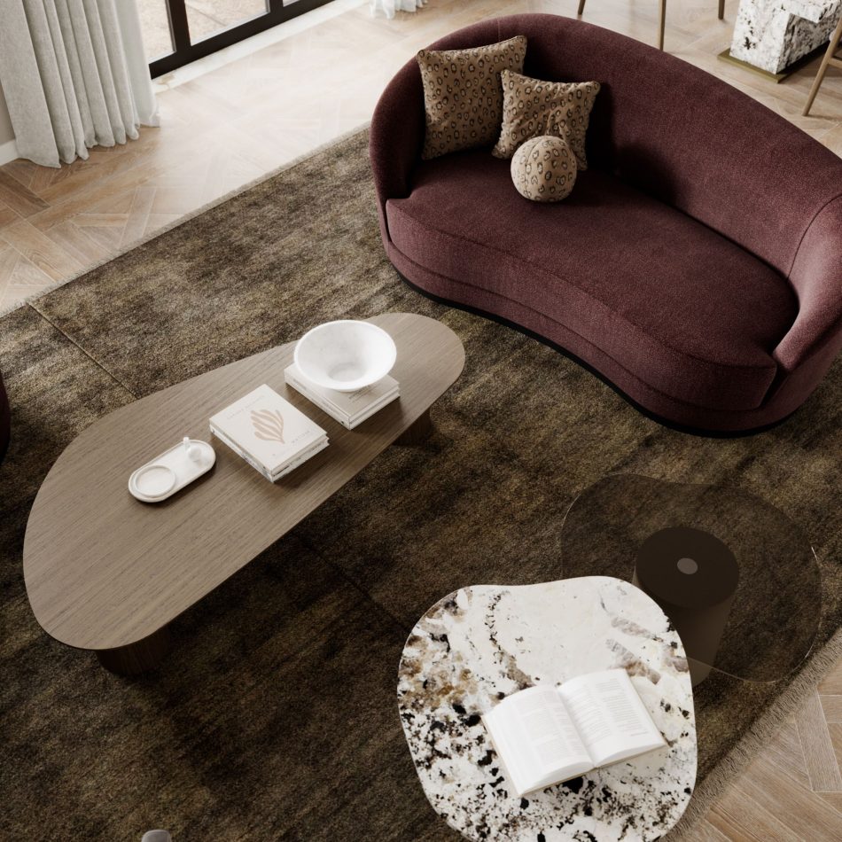 The Natur Coffee Table in a warm wood finish sits in front of a deep burgundy sofa, paired with a marbled side table and styled with books and ceramics. Soft light filters through sheer curtains, highlighting the organic silhouette of the piece. This calm setting reflects refined living room furniture, shaped by the warmth of bespoke living room design, and arranged to inspire subtle living room inspiration.