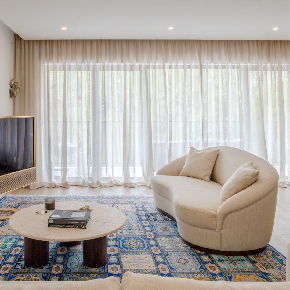 A serene living room corner featuring the Hillary Sofa, its curved lines enhancing the gentle flow of the space. The Bespoke Natur Coffee Table sits at the center with its travertine top, adding visual interest while maintaining the room’s calm identity. Soft blue accents in the rug contribute to a refined and airy environment defined by luxury furniture and natural light.