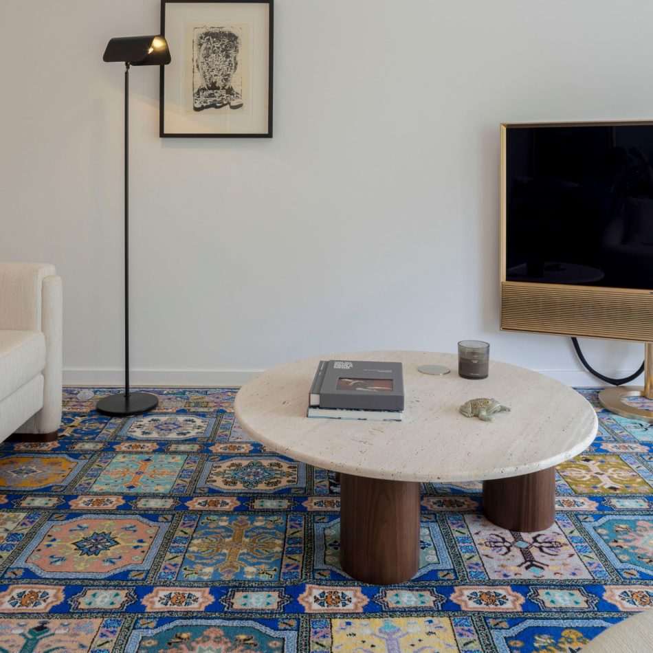 A close view of the Natur Coffee Table, where the travertine top and wooden base create a warm contrast against the patterned rug. The blue accents in the interior design add freshness without overpowering the room, supporting a balanced composition shaped by contemporary furniture and soft, natural tones.