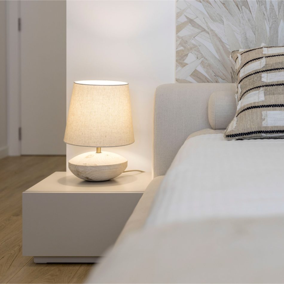 A serene bedside corner featuring the Santorini Night Table, its clean form supporting a warm, minimal lamp. The edge of the bespoke Porto Bed appears in soft neutral tones, reinforcing the room’s calm identity. Light, shadow, and subtle blue accents create a refined sense of modern bedroom design.