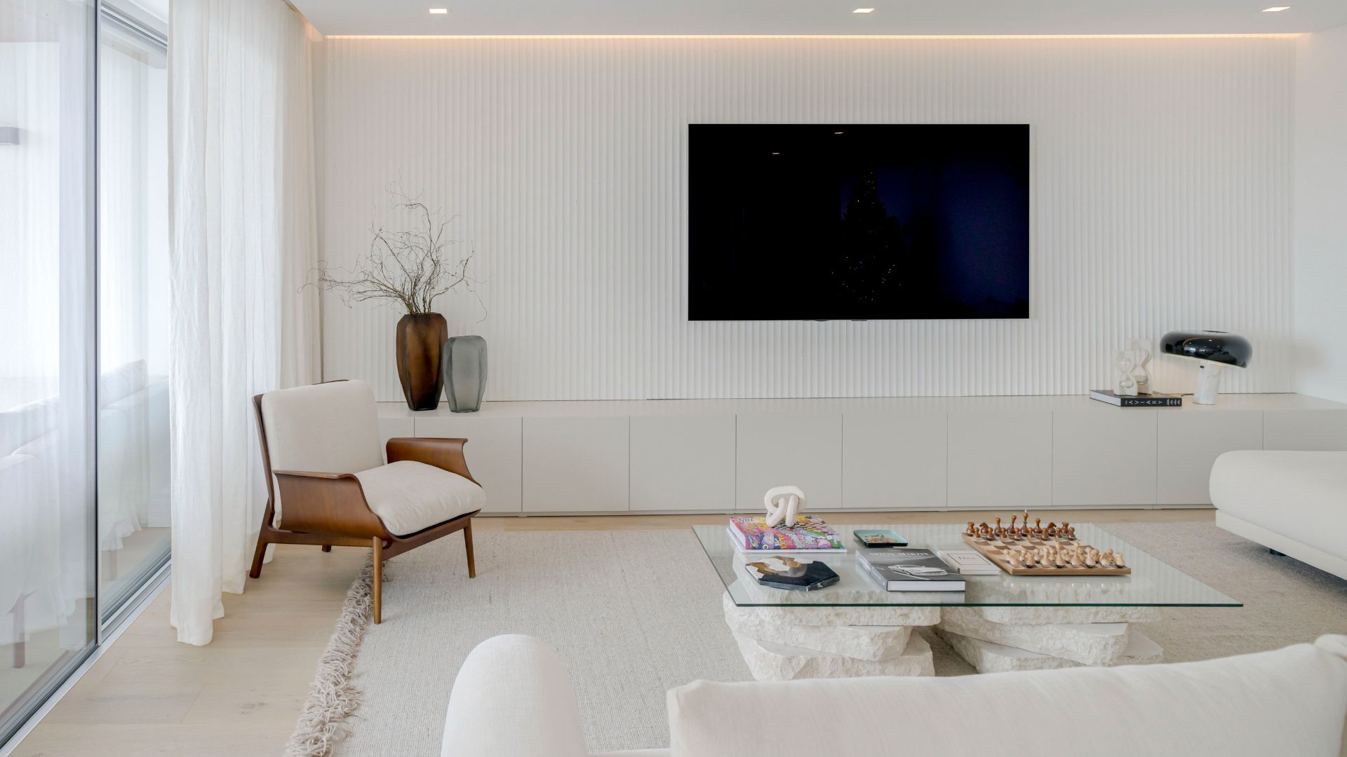 Minimalist white living room with stone coffee table, wood-accented chair, and decorative vases, emphasizing light and material simplicity.