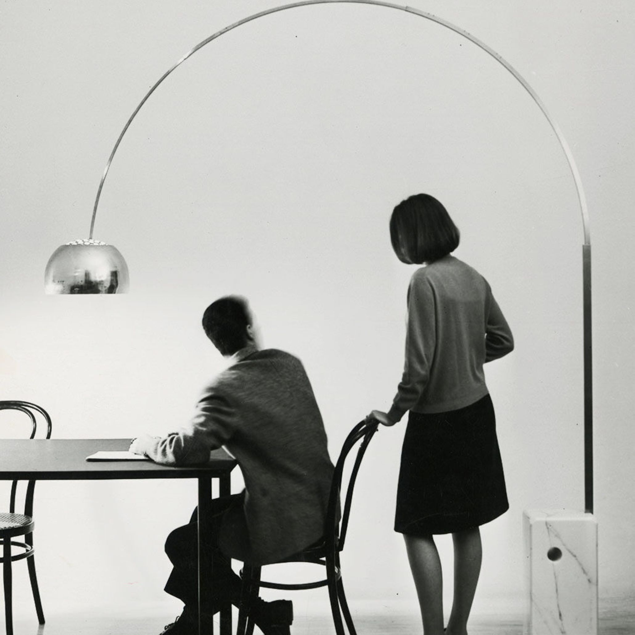 Vintage black and white photo of two people under the Arco Lamp, demonstrating its overhead reach and modernist form.