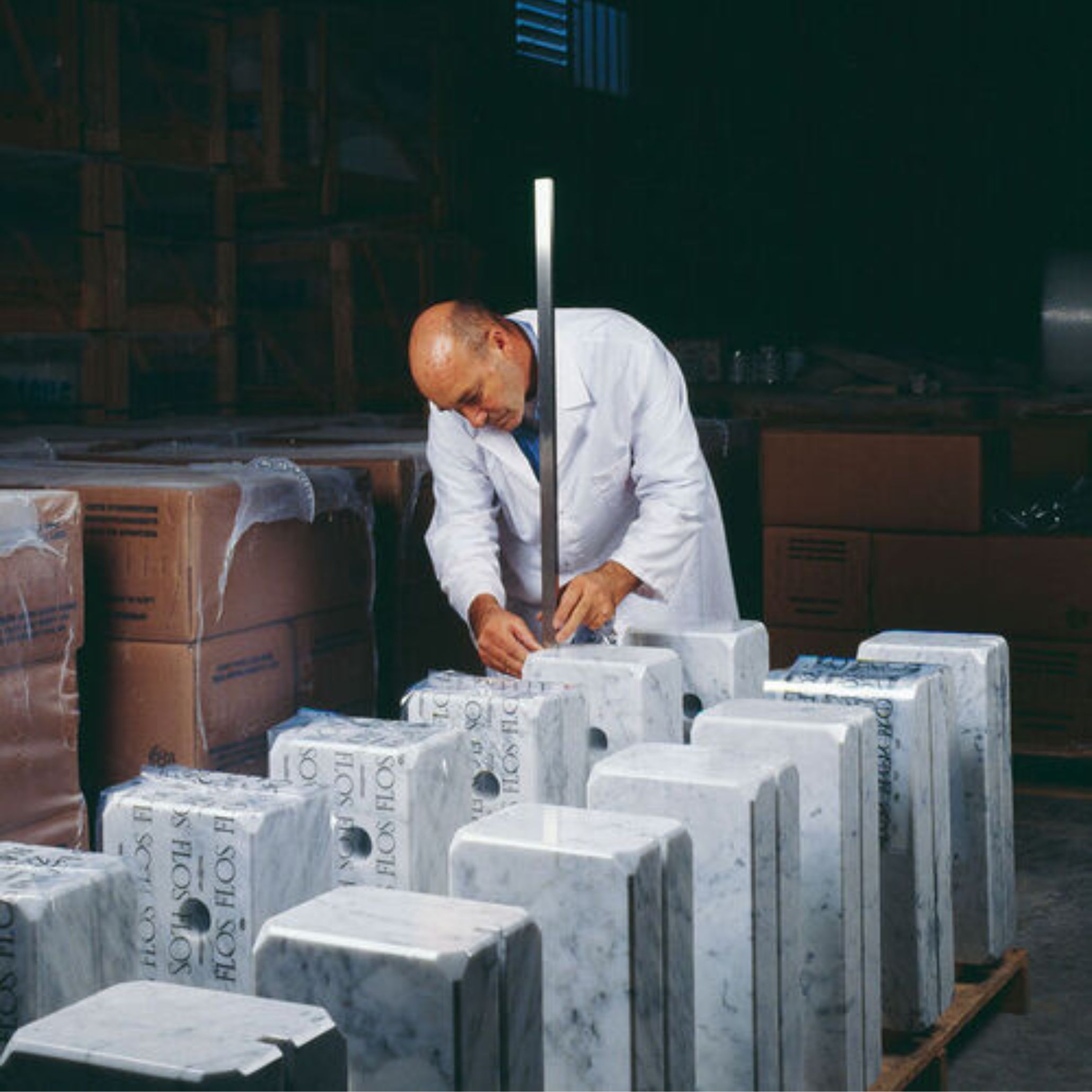 A craftsman in a white lab coat assembling marble bases for the Arco Lamp in a workshop, surrounded by multiple white Carrara marble blocks stamped with the Flos logo.