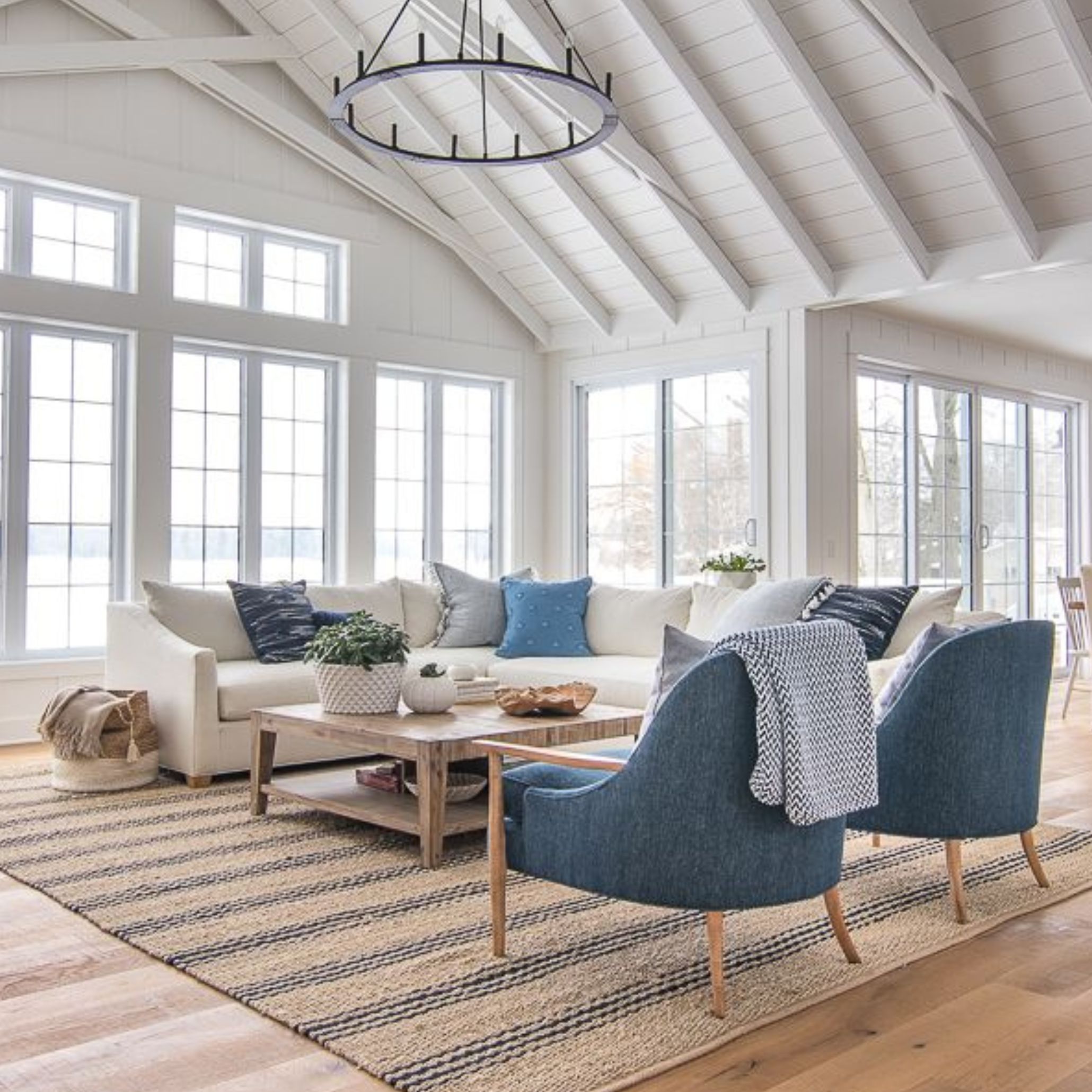A spacious and light-filled living room decorated in Coastal interior design style, with a palette of whites, beiges and blues. The space features a large sectional, elegant blue armchairs and a coastal-inspired rug, conveying the relaxed charm of Coastal Style Homes in a modern context.