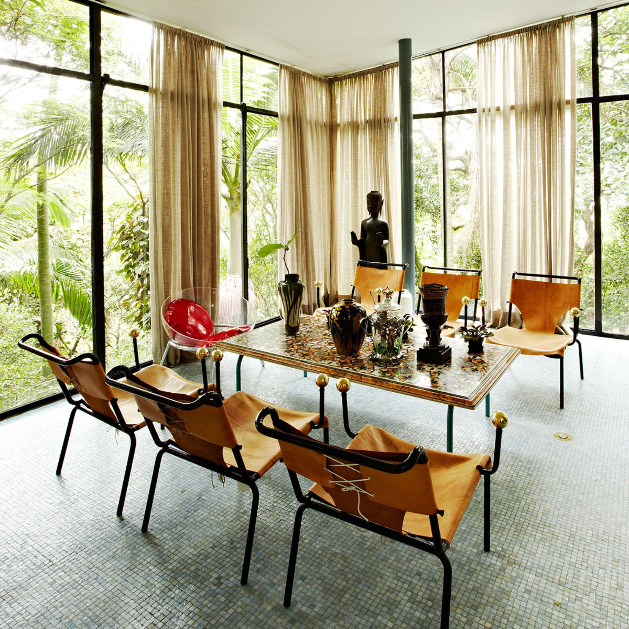Interior view of the Casa de Vidro by Lina Bo Bardi featuring a dining area with six chairs and a table. The floor-to-ceiling windows highlight her belief in spatial openness and human-centered design, central to her legacy in furniture and architectural design.