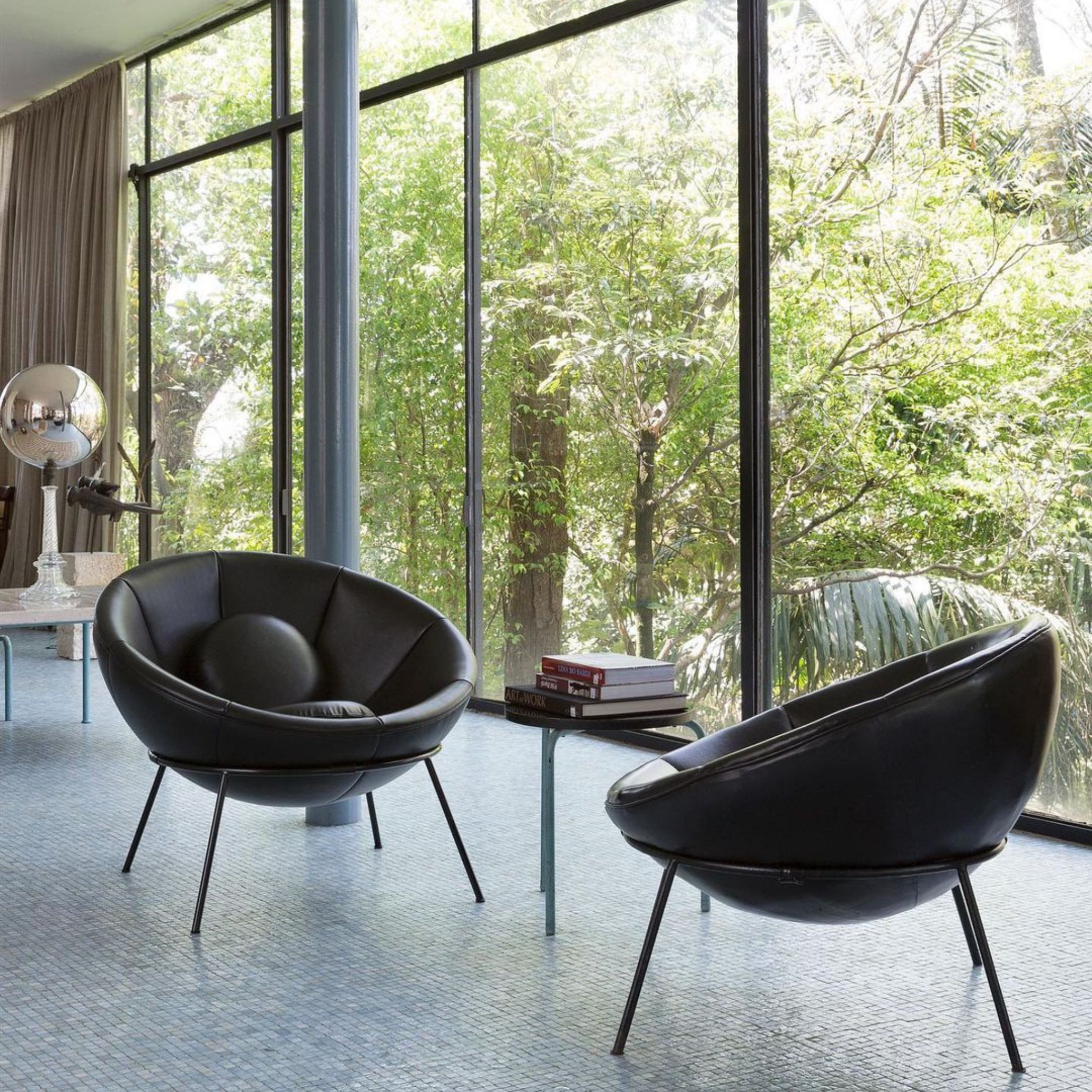 Two black leather Bardi Bowl Chairs inside Casa de Vidro, framed by expansive windows revealing lush vegetation. These pieces reflect Lina Bo Bardi’s vision of adaptable, democratic furniture design within her modern architectural spaces.