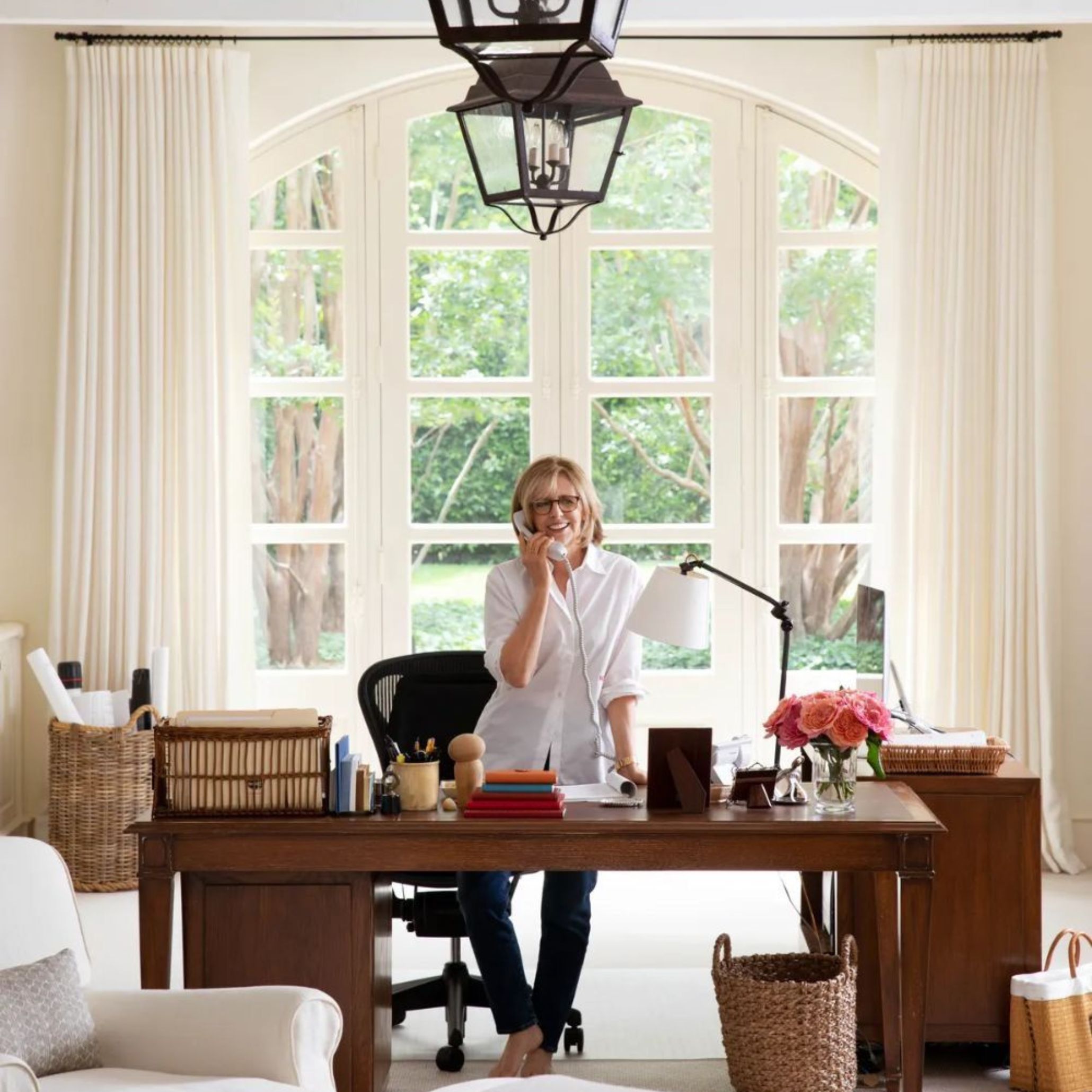 Famous director and interior style icon Nancy Meyers stands in her home office, surrounded by classic elements that define her aesthetic. The space features a traditional wooden desk, natural woven baskets, neutral drapery, and oversized arched windows that bathe the room in natural light. This image perfectly captures the understated elegance and warm, inviting ambiance that inspired the iconic Hamptons Style House and the timeless Nancy Meyers Aesthetic.