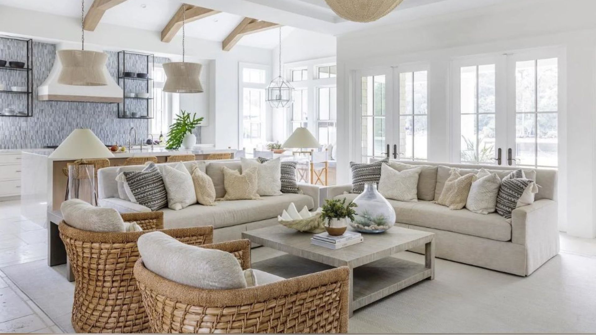A serene Hamptons Style House interior that highlights the Nancy Meyers Aesthetic through the use of warm neutrals, natural textures, and modern furniture design. The open-plan layout features a light-filled living area with woven armchairs, linen sofas in beige and off-white tones, and soft accents in grey and cream. Exposed wooden beams and coastal-inspired decor enhance the sense of quiet luxury and effortless coastal living that defines this sophisticated interior style.