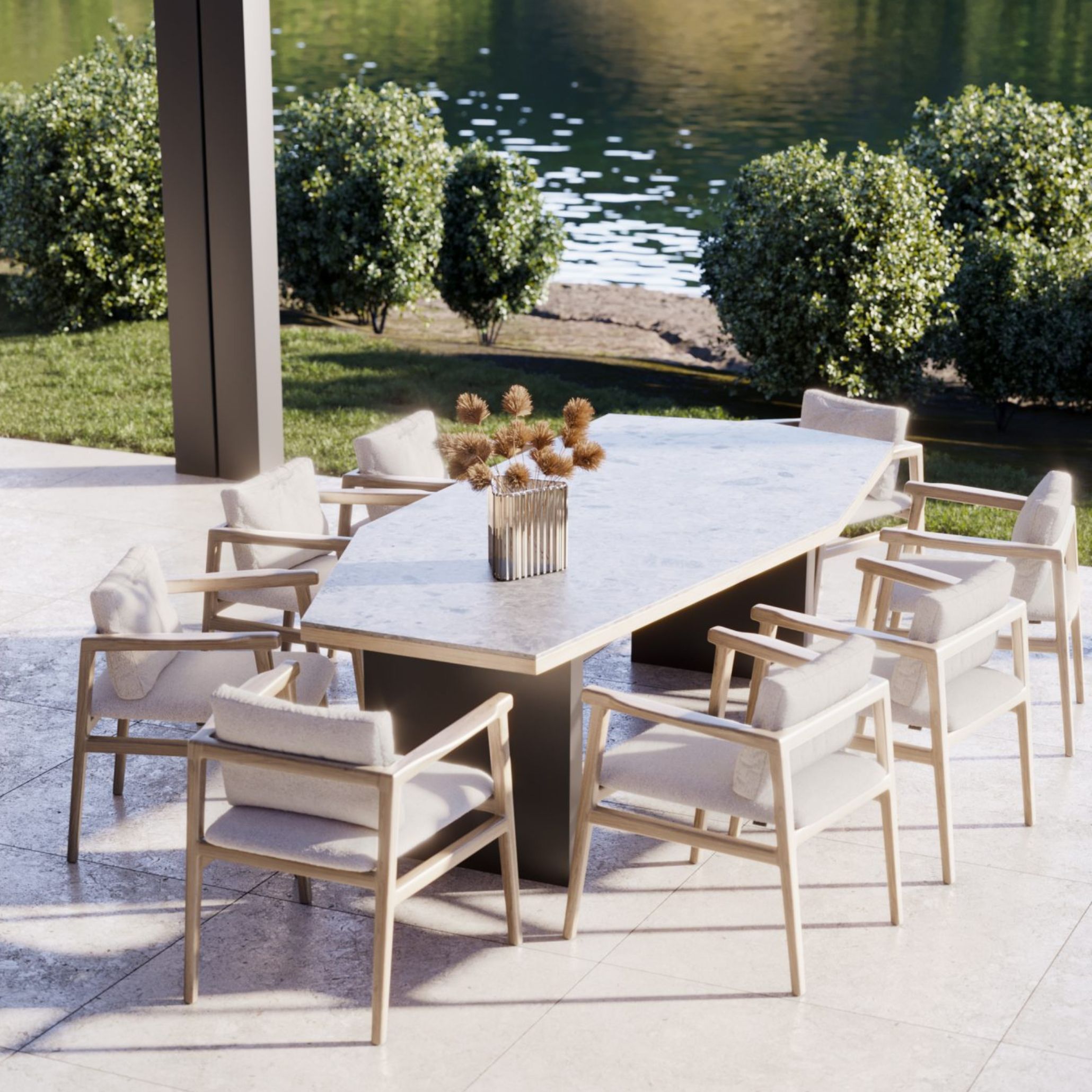 Outdoor dining area by the river, featuring the Escarpa Dining Table in a soft stone finish and eight Toro Chairs in light beige wood. The table is set with a sculptural centerpiece of dried natural elements, offering an elegant and minimalist outdoor dining table decoration with a strong connection to nature.