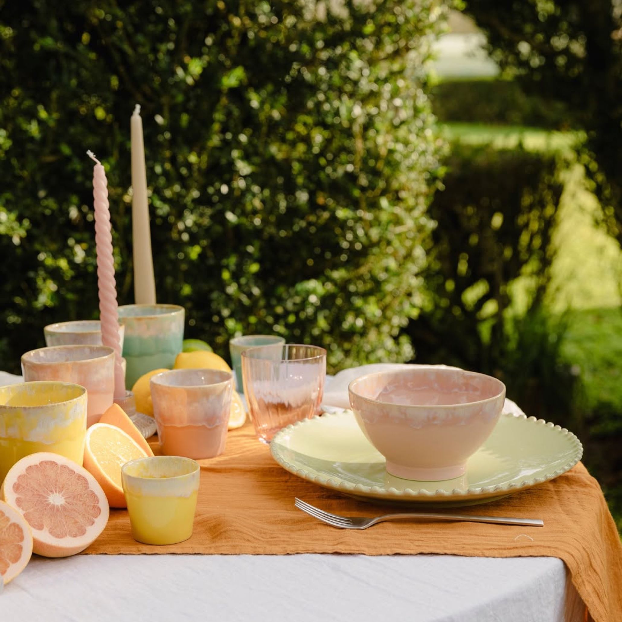 Outdoor dining table decorated with gradient-toned pastel ceramics in peach, yellow, aqua and pink. Glass tumblers, twisted candles in soft tones and fruits add vibrant charm and freshness to this colorful summer outdoor table setting.