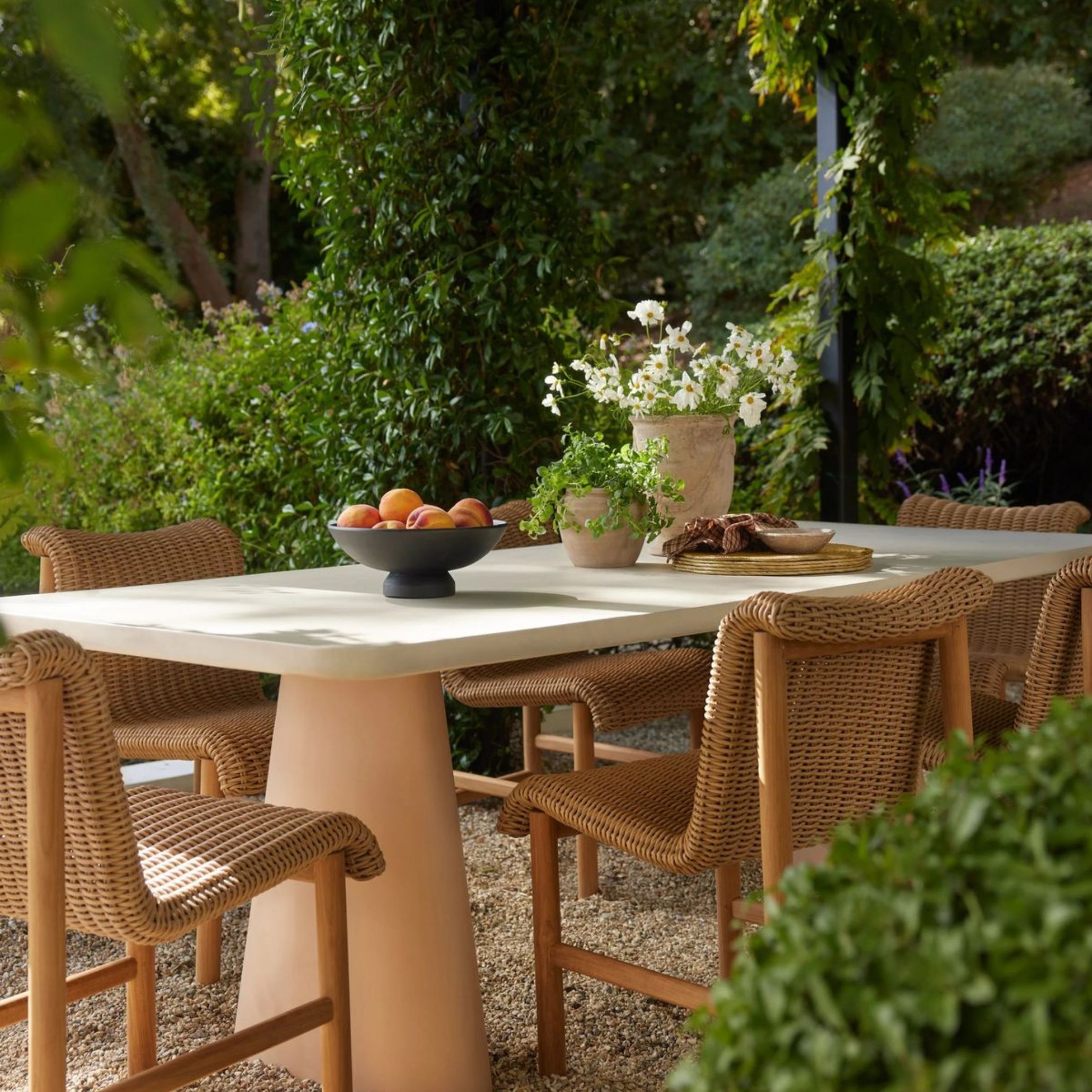 Outdoor table setting in a garden, featuring a cream ceramic table with peach-toned pedestal base, surrounded by woven rattan chairs with light wood frames. White wildflowers, potted herbs and terracotta ceramics complete the earthy, Mediterranean-inspired alfresco dining table setup.