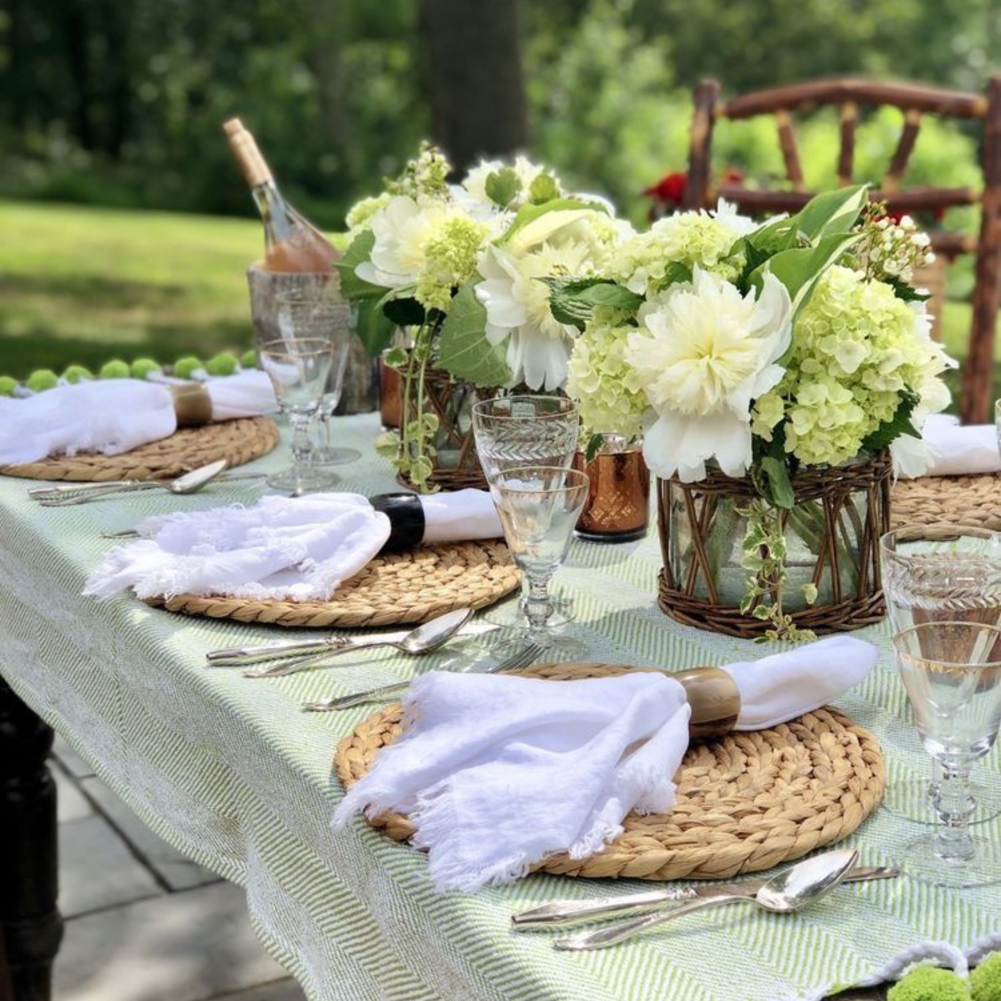 Outdoor table setting with a green herringbone cloth, round wicker placemats, white cotton napkins with fringe, and rustic wooden chairs. The centrepieces feature wicker vases filled with large white hydrangeas, dahlias and green foliage, creating a fresh and serene natural colour palette perfect for a summer outdoor dining table decoration.