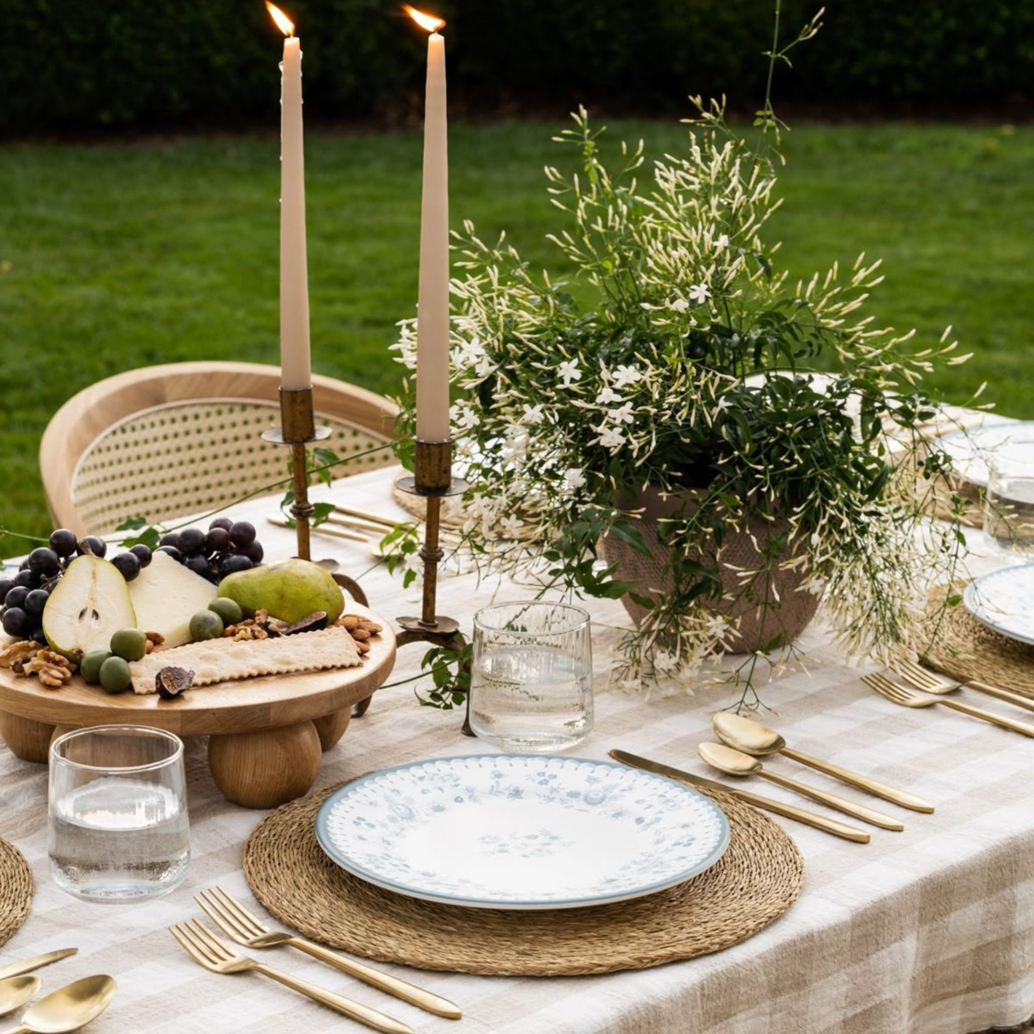 Alfresco dining table decorated with a neutral plaid tablecloth, golden cutlery, handwoven placemats, and white porcelain plates with blue floral patterns. A large arrangement of white jasmine and wild greens adds organic movement, while two tall ivory candles on brass holders bring warmth and intimacy to this outdoor dining table decoration.