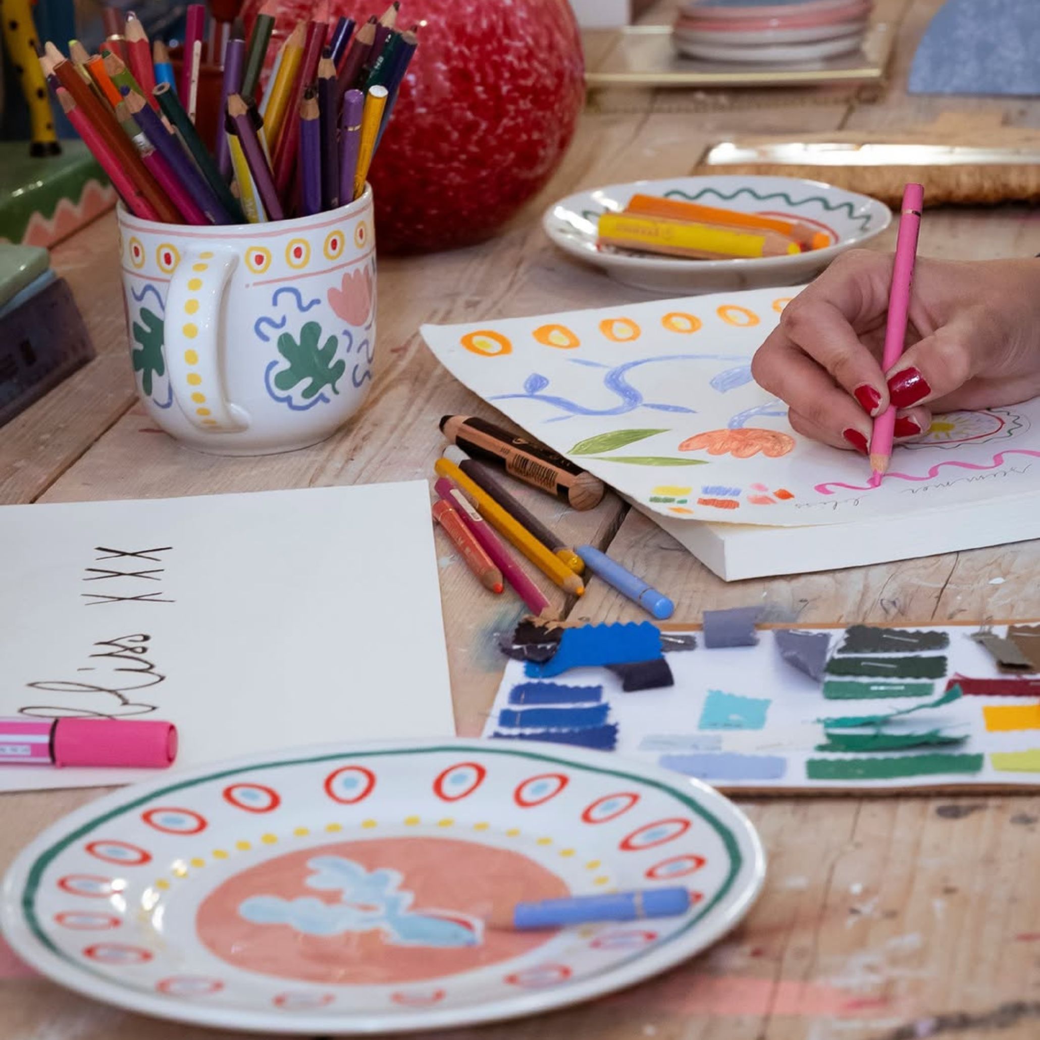 Artistic flat lay on a rustic wooden table, with hand-painted ceramic plates in pink, green and orange, surrounded by coloured pencils, crayons, and sketches of floral motifs. The mix of creativity and ceramics adds a unique focal point and elevates the outdoor dining table decoration with personality and charm.