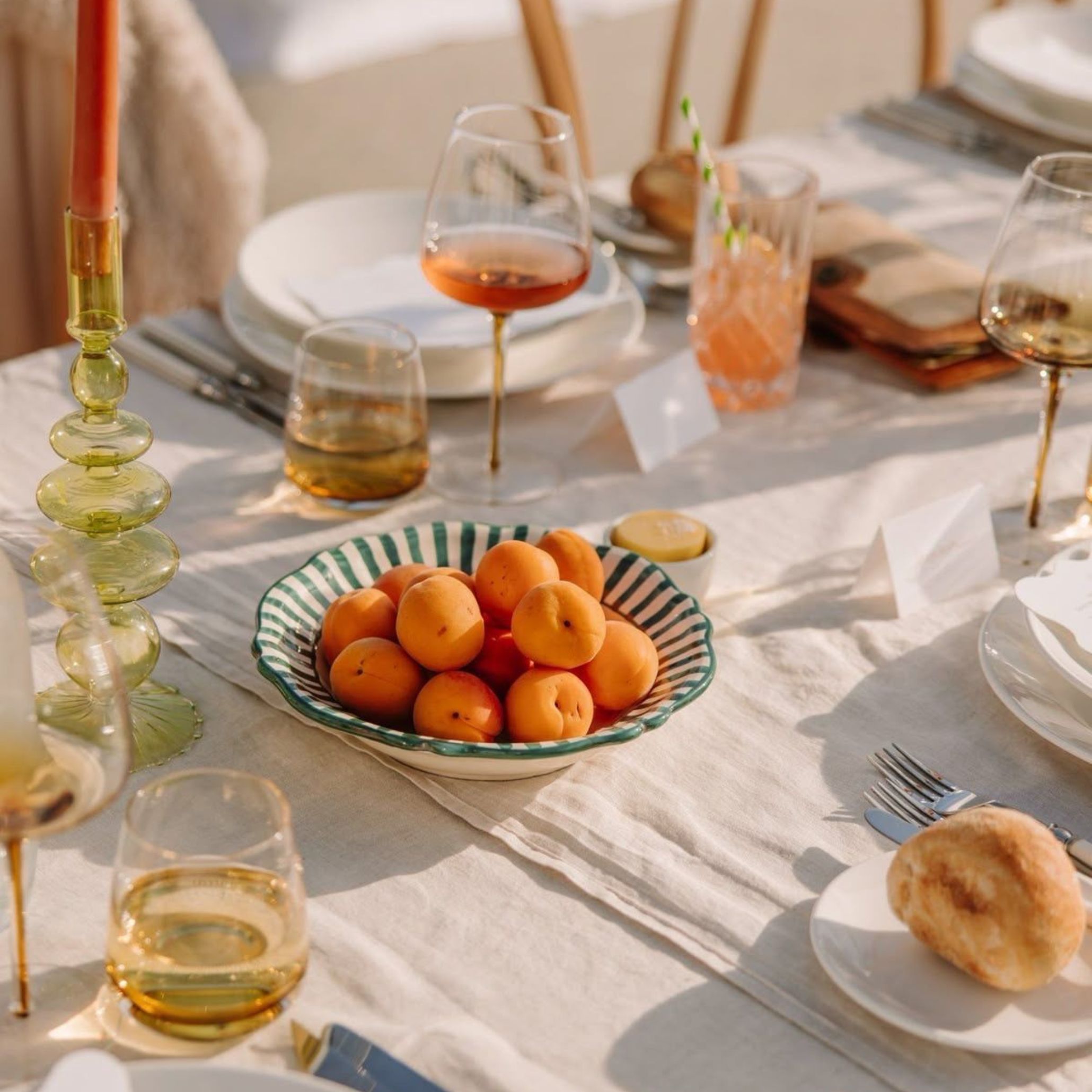 Outdoor table setting featuring a striped ceramic bowl filled with ripe apricots, used as a natural and colourful centerpiece. Surrounded by golden wine glasses, green glass candle holders, and soft linen napkins, this composition brings a fresh and effortless elegance to the alfresco dining table decoration.