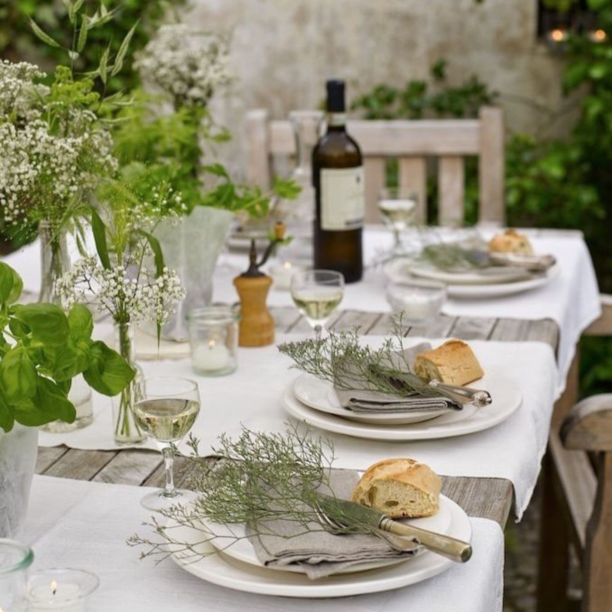 Elegant yet minimal outdoor table decorated with a white linen tablecloth and wooden furniture. Each plate is paired with a grey linen napkin, topped with sprigs of fennel or dill, a small rustic bread roll, and simple cutlery. Glasses of white wine and wildflowers in jars complete this intimate and subtle alfresco dining table setup.