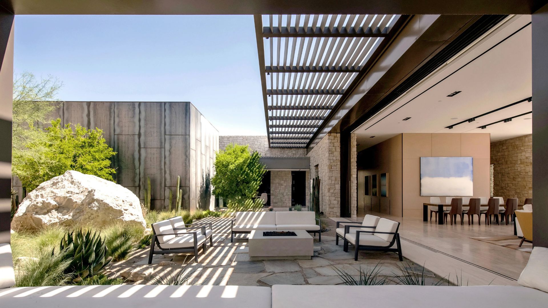 Minimalist white lounge chairs surround a geometric fire pit on a desert-inspired terrace. The scene includes natural stone walls, warm wooden pergolas, and sculptural desert plants, embodying the 2025 trend of unified living spaces that blur the boundaries between interior and exterior comfort.