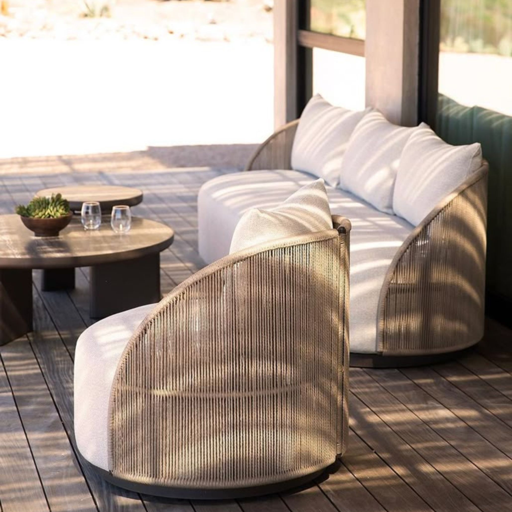 Two round outdoor armchairs with woven rope backs and plush cream cushions sit on a wooden deck bathed in dappled sunlight. With tactile surfaces, natural fibers, and soothing forms, this setup reflects a sustainable, nature-embracing approach to furniture design.
