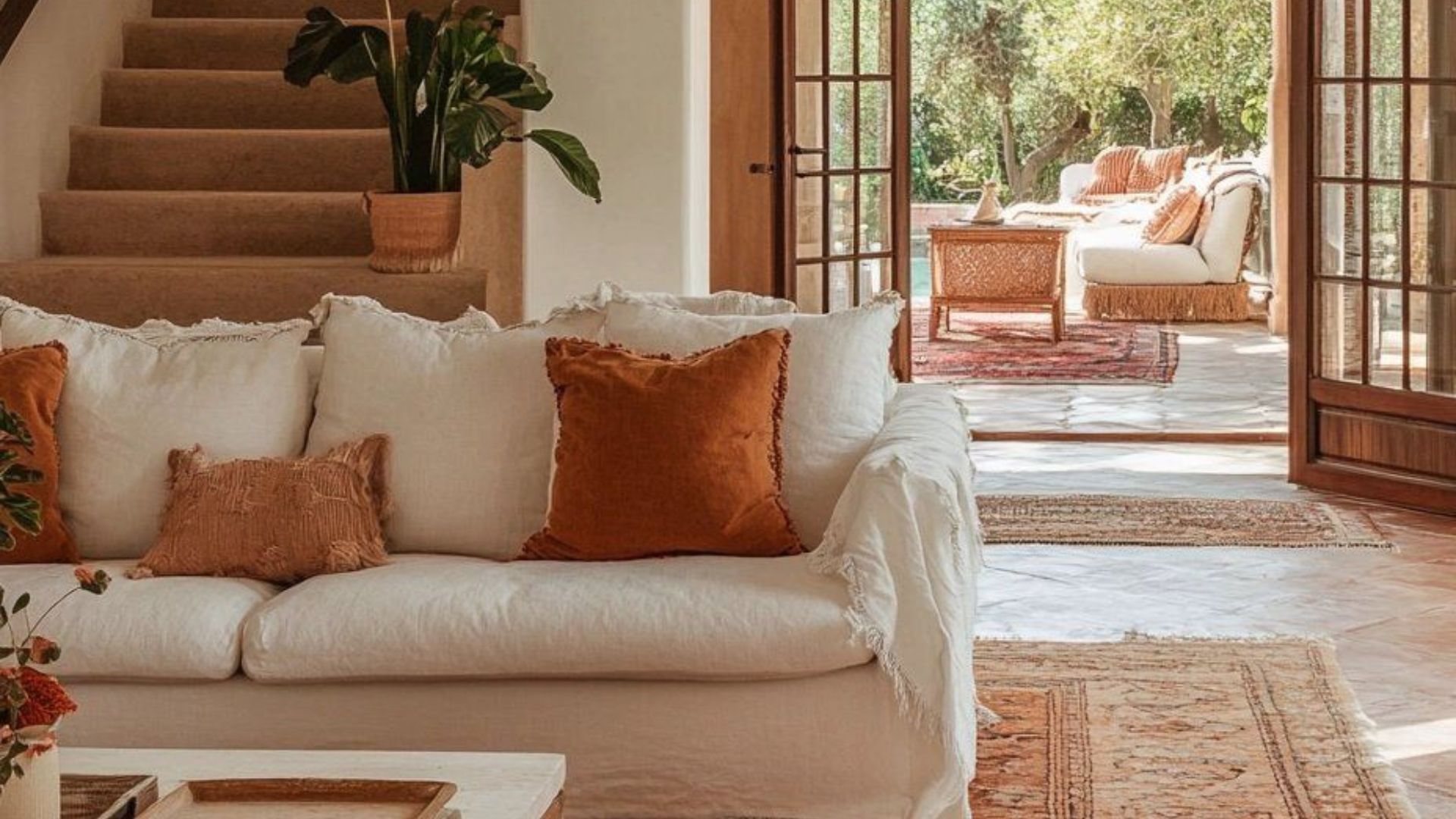 Elegant living space blending interior and exterior seamlessly, with a white linen sofa, terracotta pillows, rustic tiled flooring, and wooden ceiling beams. Outside, a matching lounge continues the aesthetic, showing the Tuscan approach to cohesive, nature-inspired spaces using an earthy, sun-kissed palette.