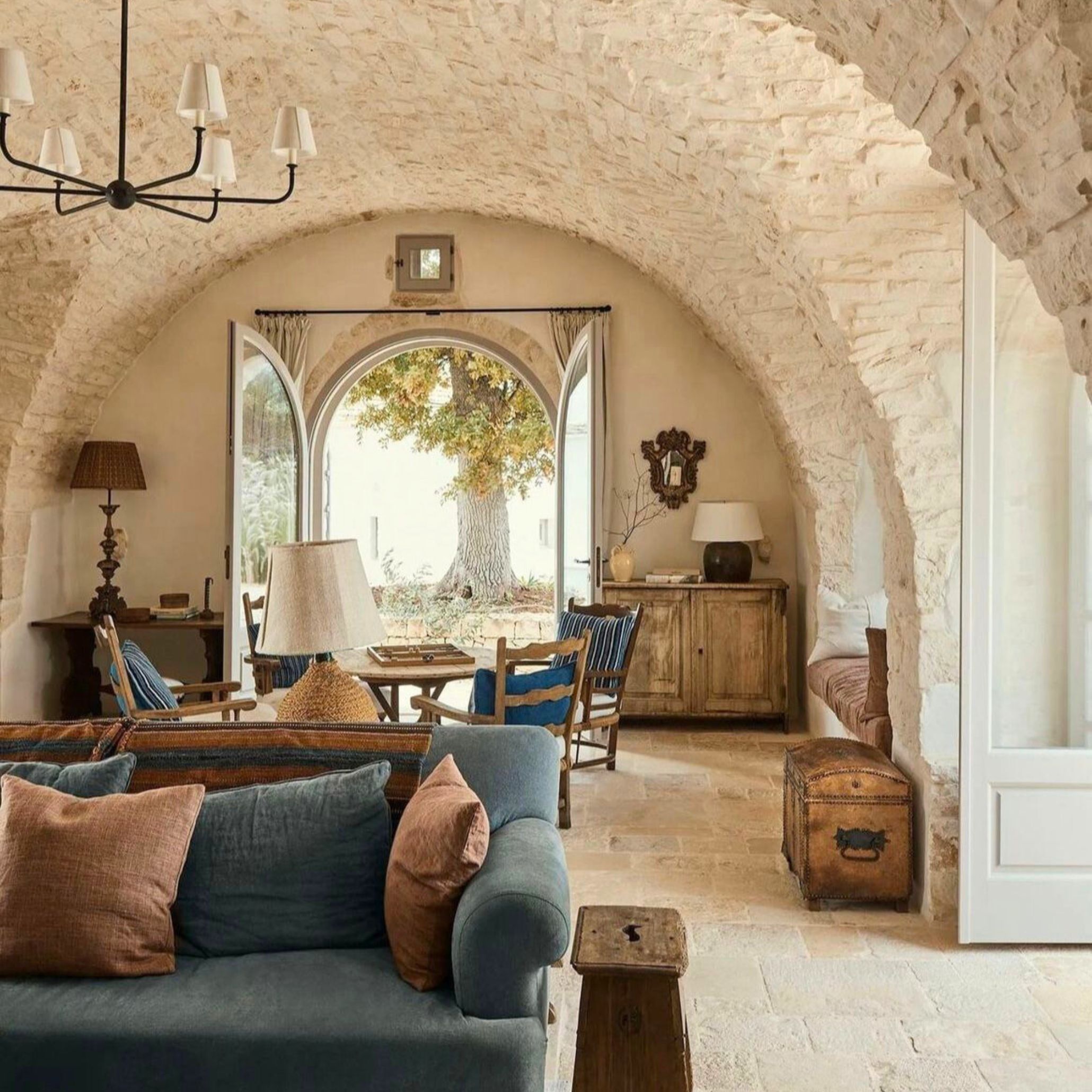 Expansive Tuscan open-concept living space framed by grand arched stone walls, with carefully placed wood furniture, woven fabrics in muted oranges and blues, and iron accents. This interior demonstrates the textural richness and raw beauty of natural materials essential to Tuscan interior design.