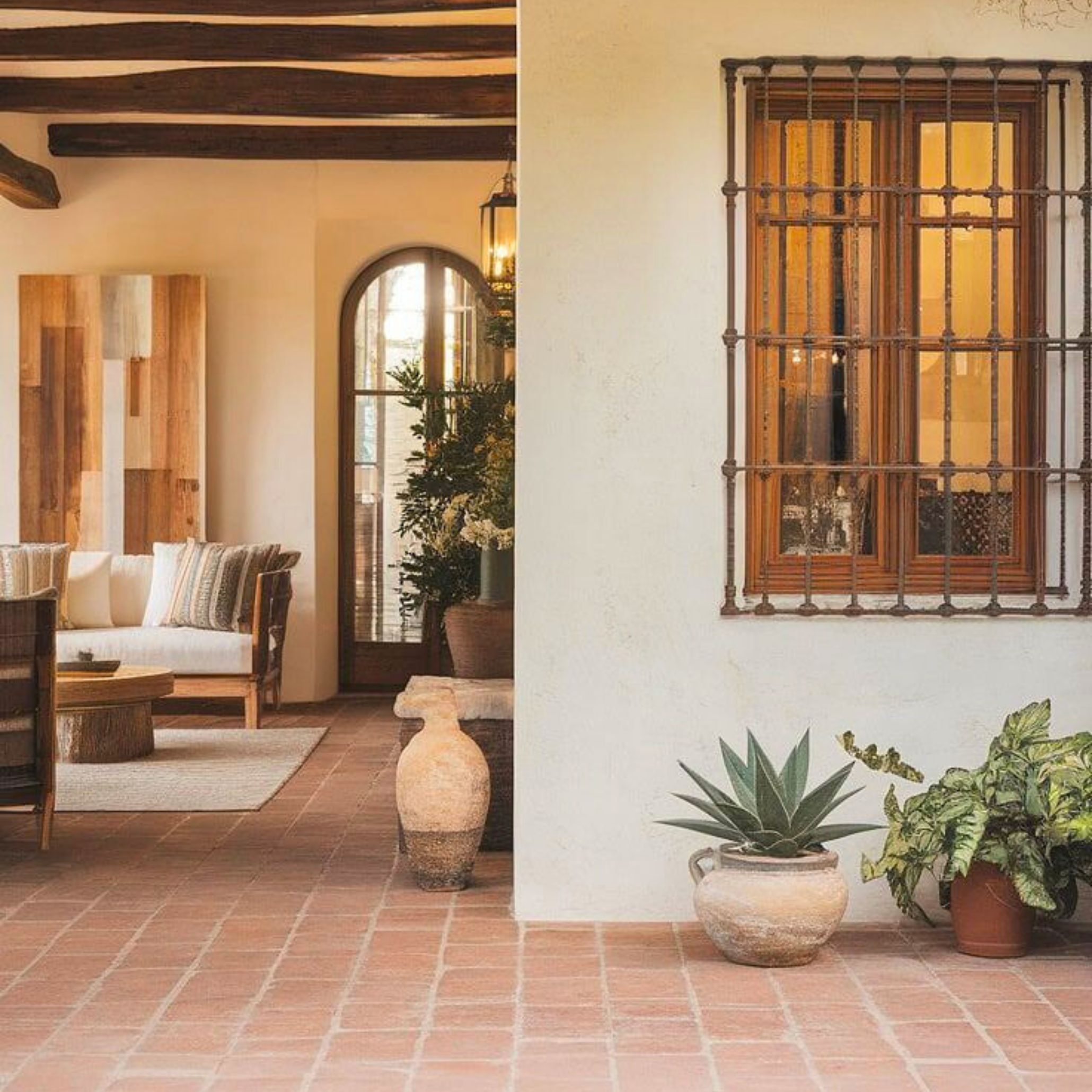 Charming Tuscan-style courtyard featuring terracotta floor tiles, rustic wooden ceiling beams, and a neutral-toned seating area with textured cushions. The arched doorway and iron window grille add architectural authenticity, while clay pots and potted greenery enhance the organic, earthy palette. This space exemplifies Tuscan interior design’s signature use of natural materials, handcrafted details, and warm, welcoming ambiance.