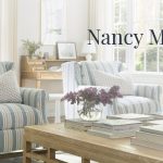 Cozy and elegant living room inspired by the Nancy Meyers aesthetic, featuring two striped armchairs with patterned cushions, a wooden coffee table stacked with books, fresh lilac flowers in a glass vase, and large windows flooding the room with natural light.