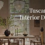 Cozy Tuscan-style dining room showcasing rich wooden furniture including a large dining table and chairs, illuminated by two classic chandeliers hanging from a rustic ceiling. Floor-to-ceiling windows reveal a serene outdoor landscape, flooding the space with natural light and enhancing the warm, inviting ambiance of traditional Tuscan interior design.