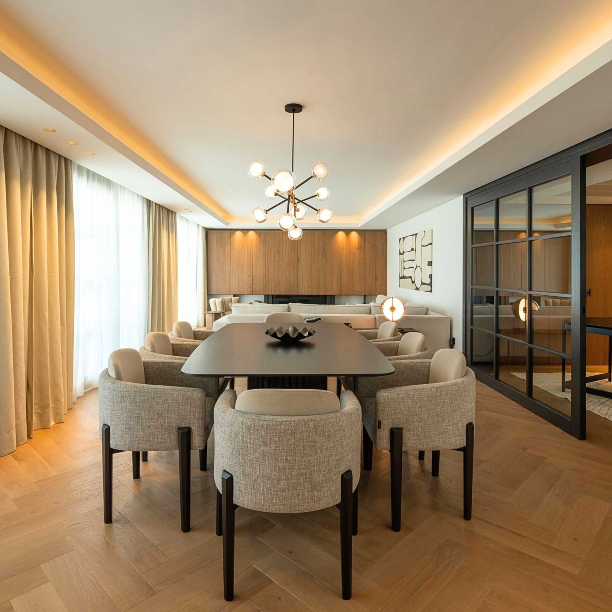 The dining area of A Polished Edge villa features His II Chairs around a sleek dark table, framed by warm wood tones and soft lighting. A sculptural chandelier and herringbone flooring enhance the sense of modern sophistication. The space reflects fluid transitions and a refined balance between elegance and practicality.