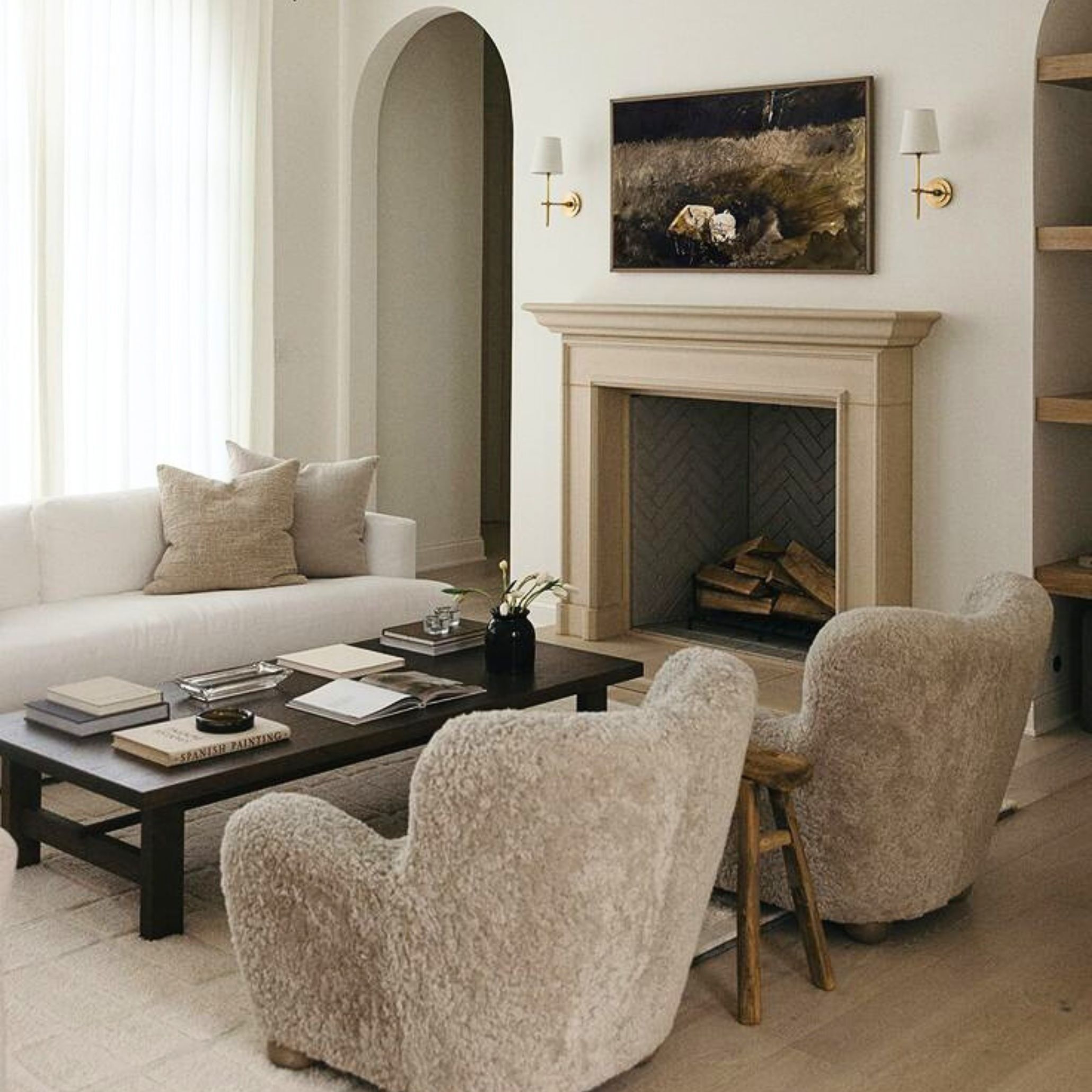Cozy Transitional living room with a beige fireplace, pastoral painting, gold sconces, plush armchairs, and a dark wood coffee table - highlighting the balance of classic charm and modern simplicity in a elegant setting.