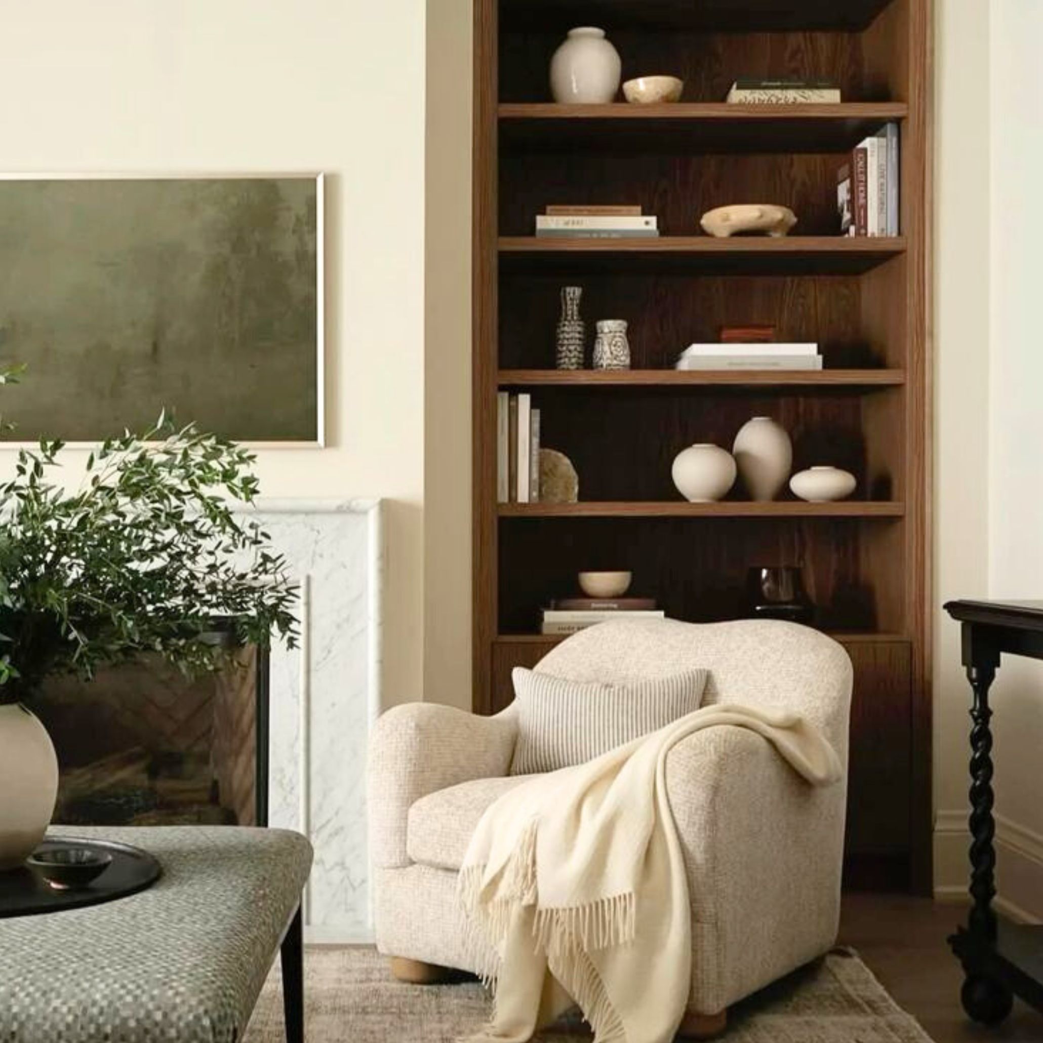 Cozy living room corner with a textured cream armchair, built-in wooden bookshelf, and marble fireplace. Showcasing Transitional Design’s layered warmth, classic influences, and serene composition.