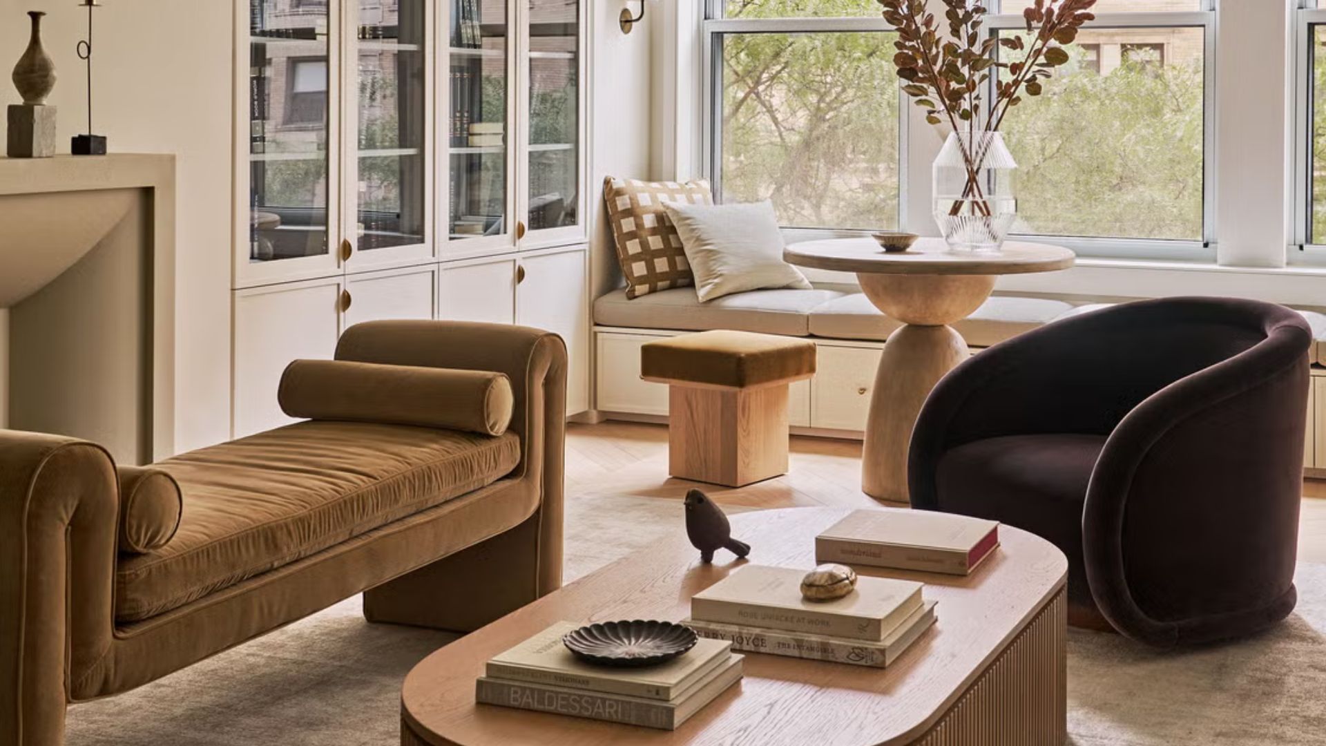 A warm, thoughtfully styled living room featuring a brown velvet chaise, a curved dark armchair, and a light wood coffee table layered with books and decorative objects. Soft natural light fills the space through large windows, highlighting the built‑in window seat, glass‑front cabinet, and sculptural accents that reflect the 2026 interior design focus on warmth, texture, and lived‑in elegance.