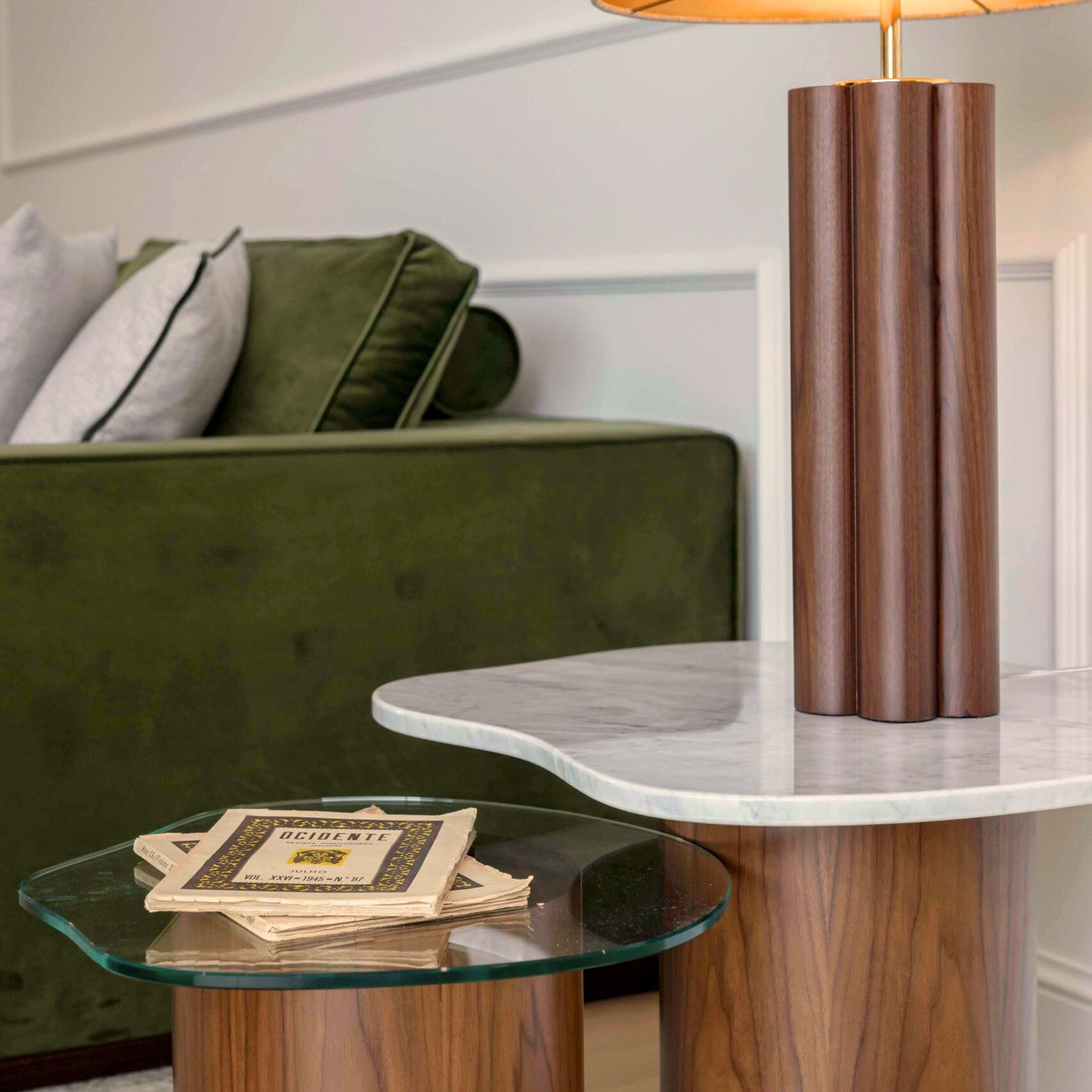 A warm, inviting corner featuring a green velvet sofa layered with soft gray cushions, paired with two Natur side tables in marble and glass. A sculptural wooden lamp casts a gentle glow over a vintage booklet, adding a touch of personal history. The mix of textures and collected objects reflects the essence of lived‑in spaces, where comfort, character, and quiet memories shape the atmosphere.