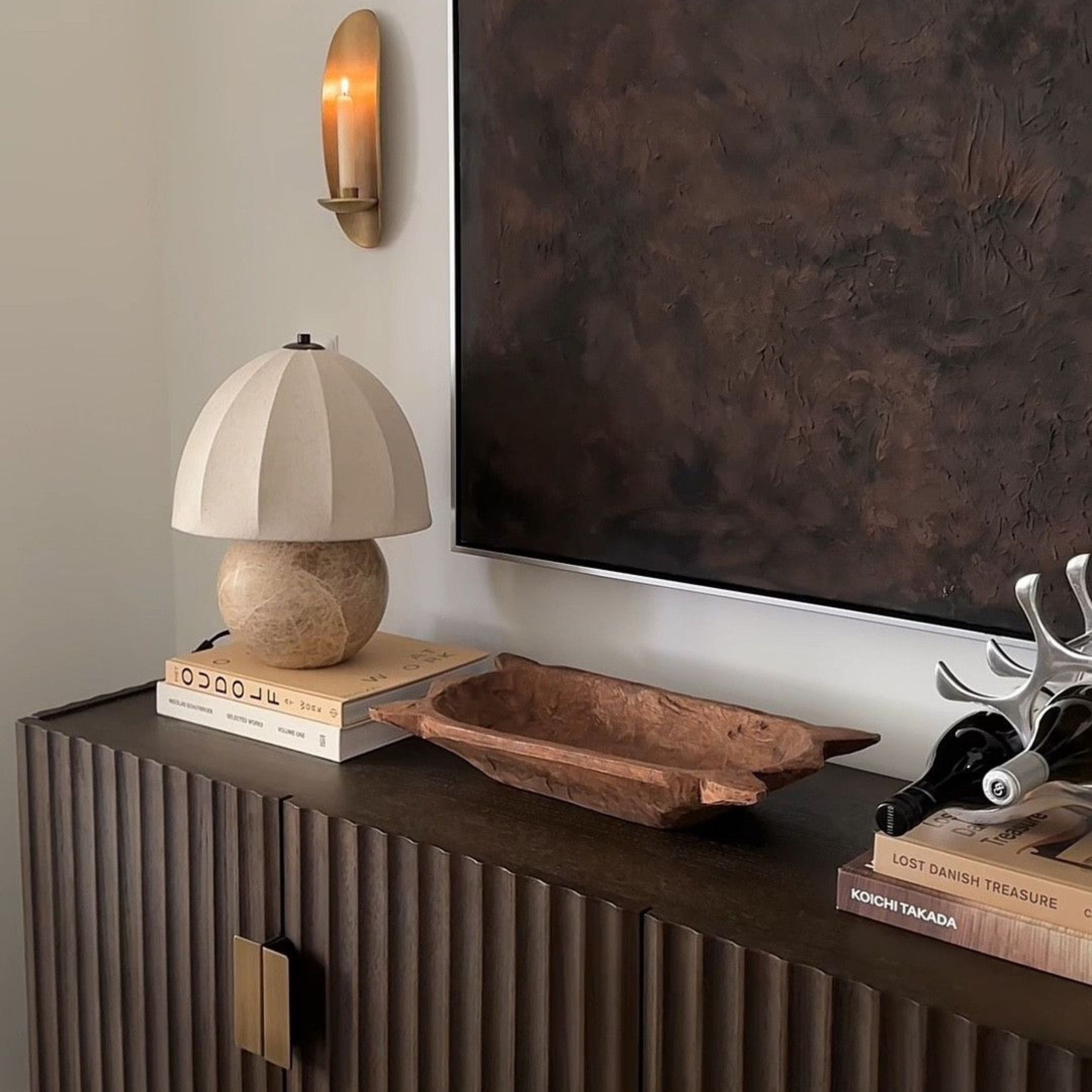 A modern vignette featuring a dark wood cabinet with vertical grooves, topped with sculptural accents - a textured lamp, carved bowl, metallic wine holder, and stacked books. A large abstract painting and brass wall sconce complete the scene, showcasing the 2026 emphasis on craftsmanship and decorative detail. Each element adds texture and personality, turning a functional corner into a refined visual statement.
