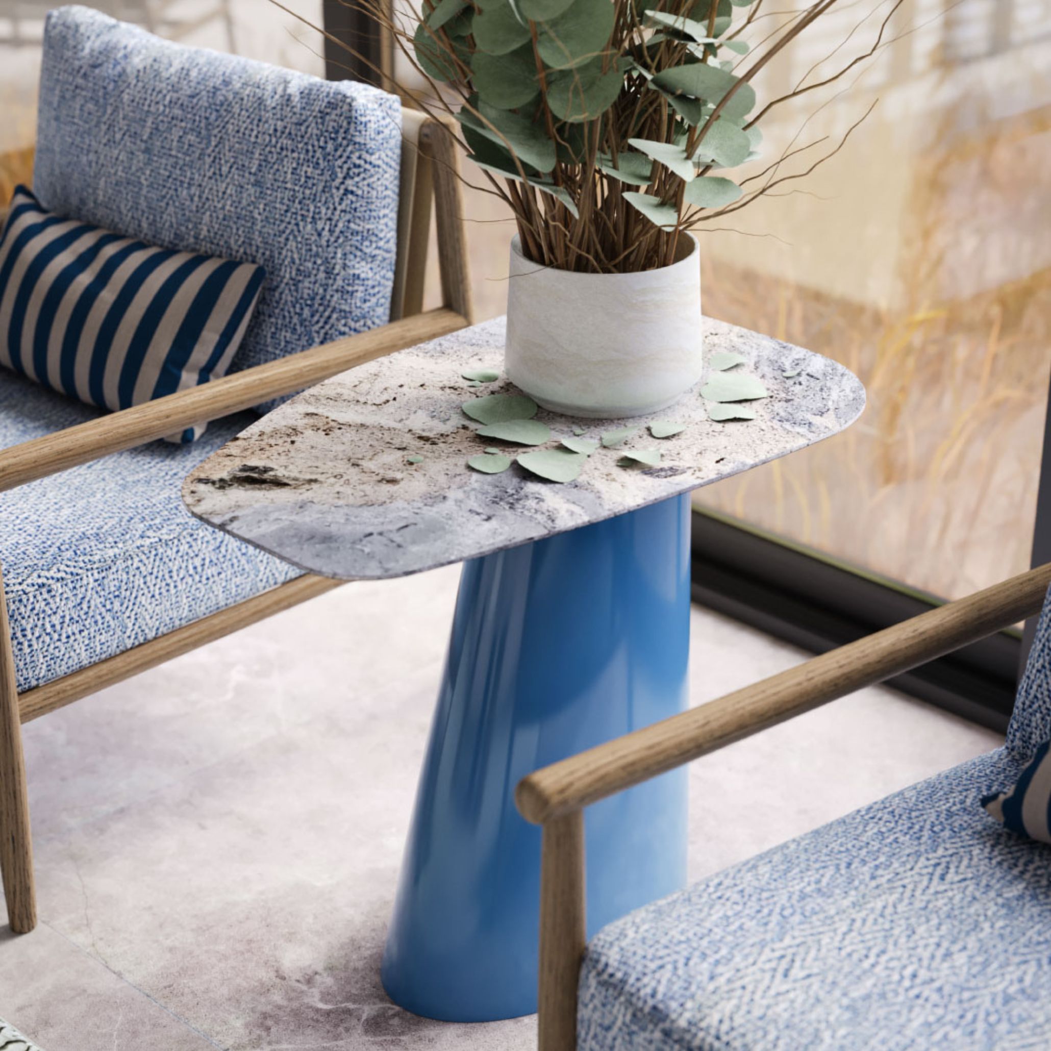 Two textured armchairs sit beside the Stone Side Table, finished in glossy blue lacquer with a sculptural base and stone-textured top. A ceramic vase with eucalyptus adds softness, while the lacquered surface introduces clarity and modern contrast - capturing the 2026 trend of luminous materials that elevate simple forms.
