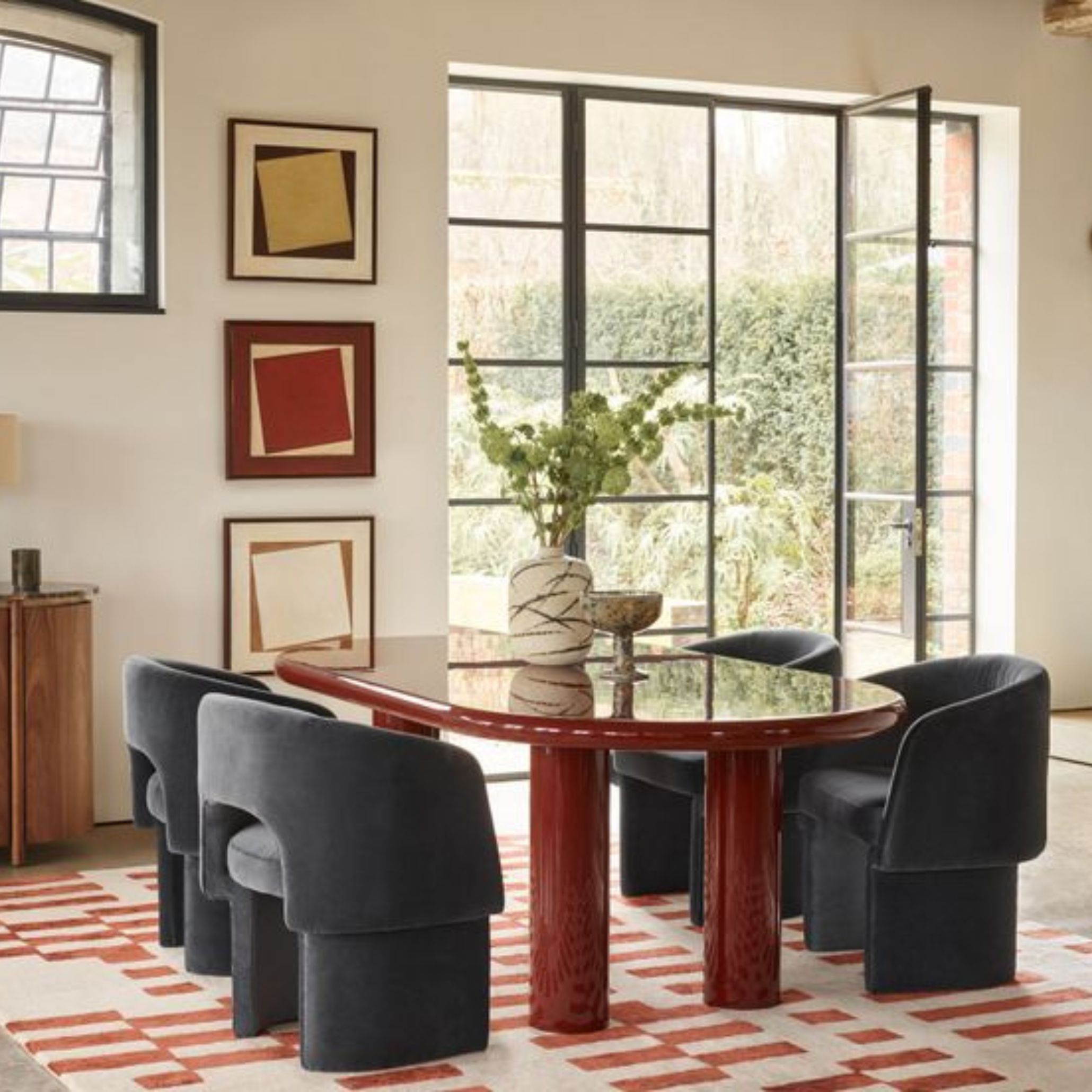 A glossy red lacquered dining table anchors a modern room, surrounded by plush dark blue chairs and framed by natural light. The table’s smooth, reflective surface adds sophistication and visual impact, illustrating how high-gloss lacquer defines 2026 interiors with clarity and contemporary edge.