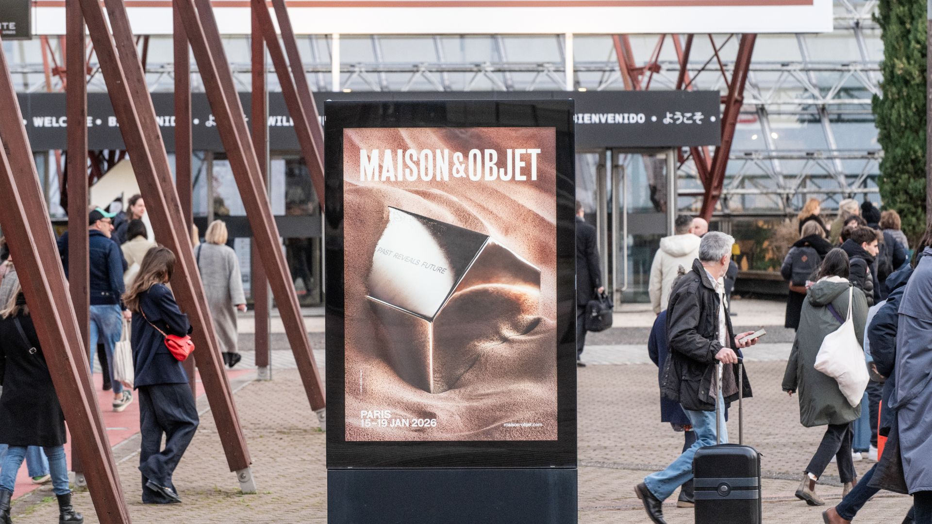 Entrance to Maison & Objet 2026 in Paris, with attendees arriving at the venue. A prominent physical installation in the foreground features the event’s theme 'Past Reveals Future' engraved on a metallic cube half-buried in sand, symbolizing the fusion of heritage and innovation in interior design.